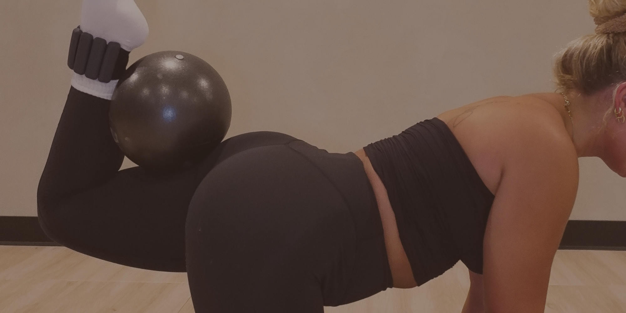 A woman with long blonde hair doing a boat pose yoga position on a black mat in a group yoga class, with other participants in the background at 8th State Hot Yoga studio.