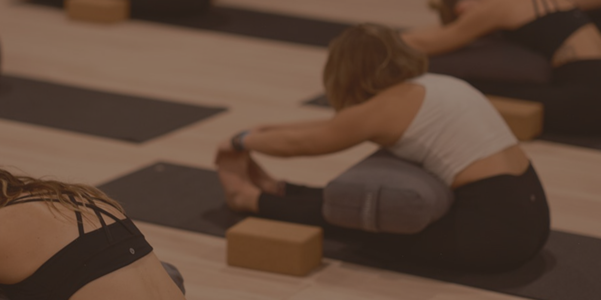 Women participating in a yoga class sitting on mats in a studio, stretching forward with their arms extended, using yoga blocks for support.
