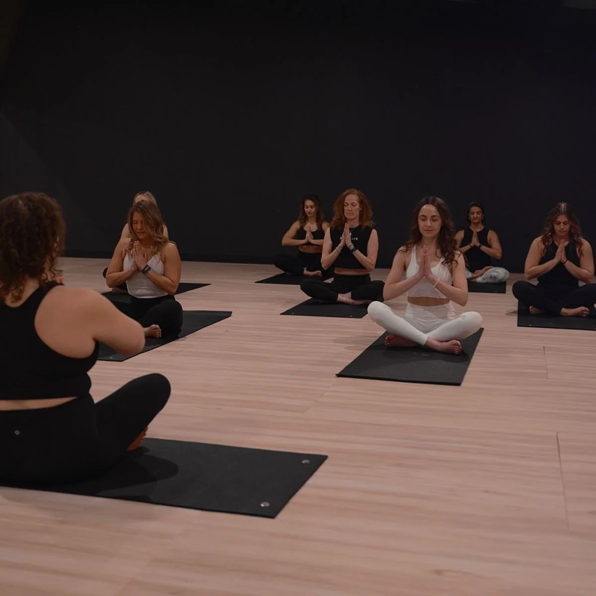 A group of women participating in a yoga class, sitting cross-legged on black mats in a meditation pose with their hands in prayer position, in a room with a dark wall and wooden floor.