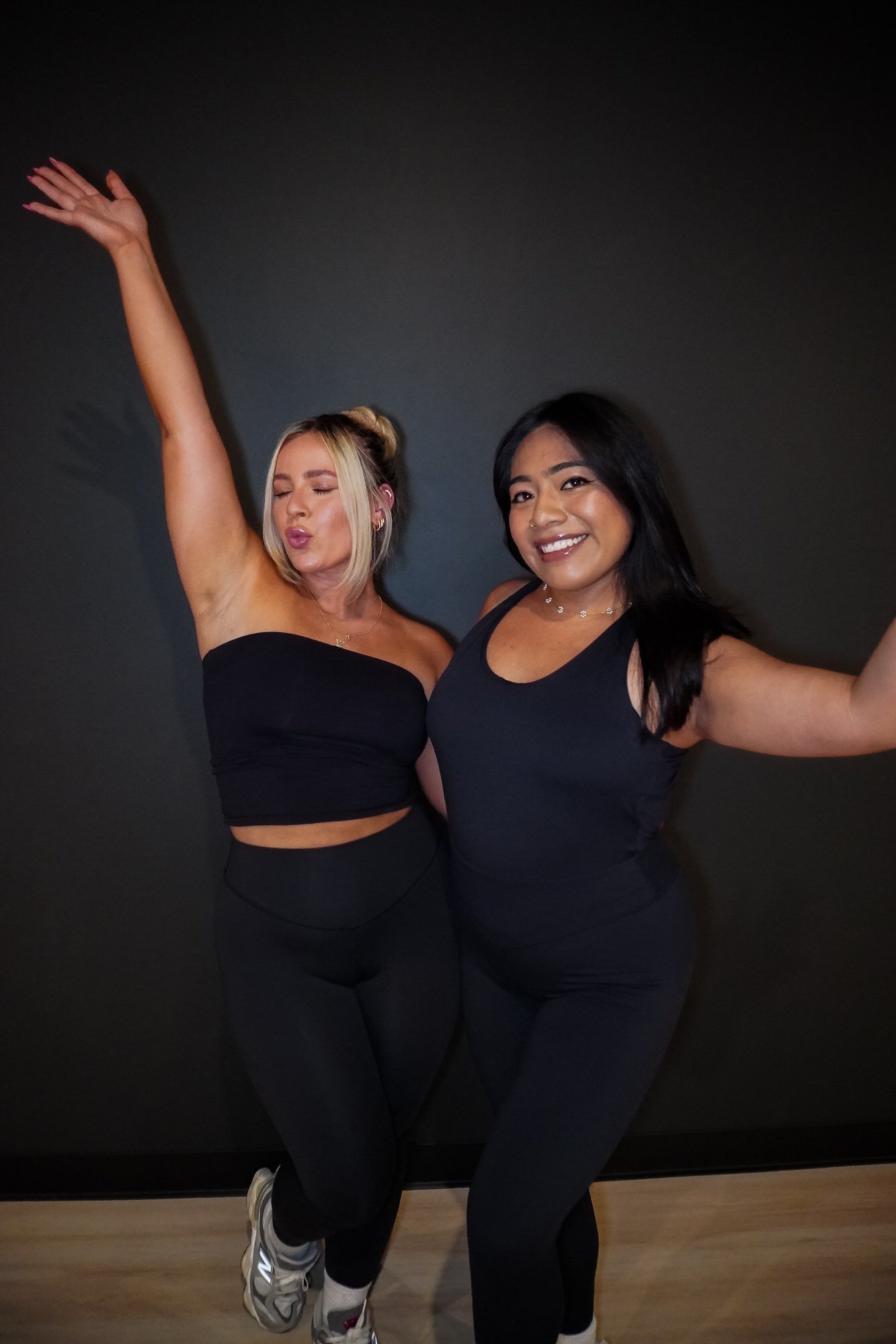 Two women in black workout clothes posing together, one with her arm raised and the other smiling broadly, against a black wall at 8th State Hot Yoga studio.
