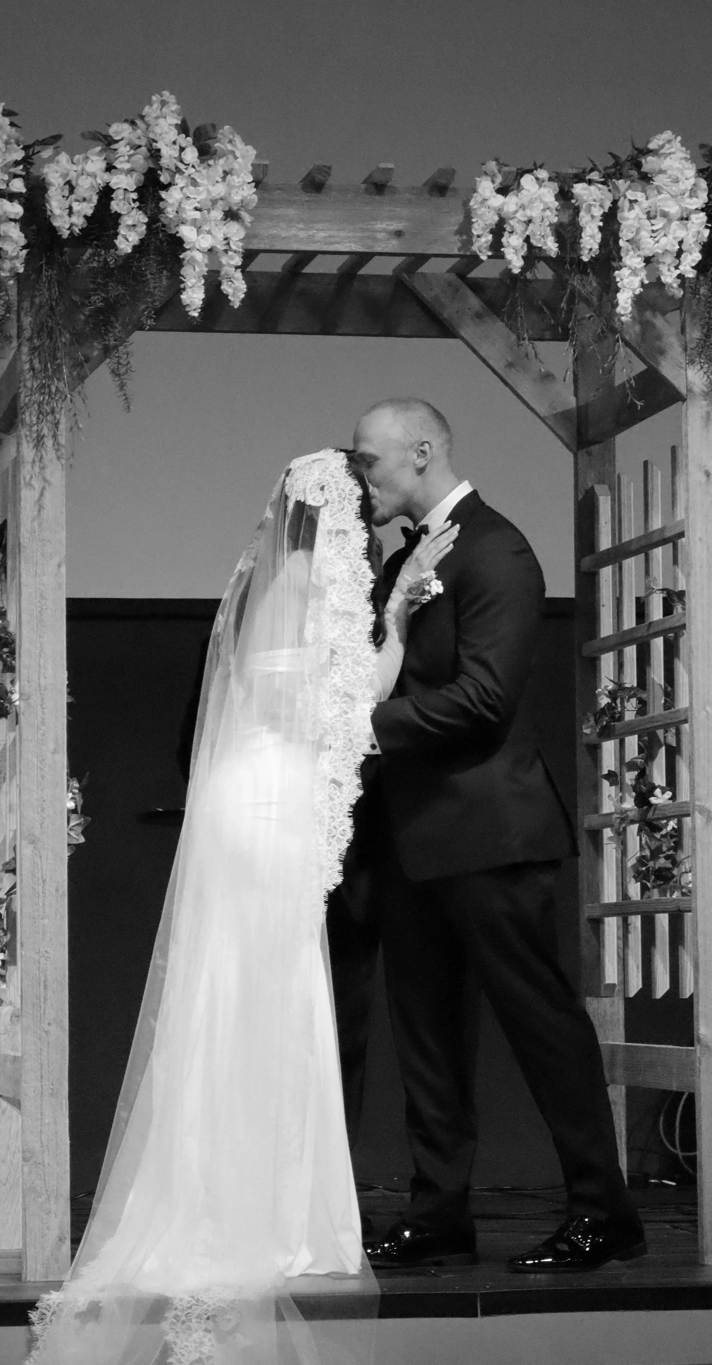 A black-and-white photograph of a wedding ceremony showing a bride and groom sharing a kiss under a decorated wooden arch.