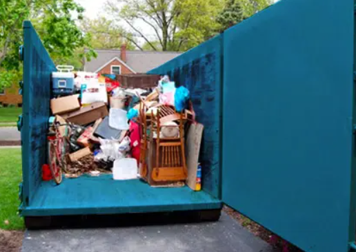 Overflowing dumpster filled with trash, cardboard boxes, furniture, and household items outdoors.