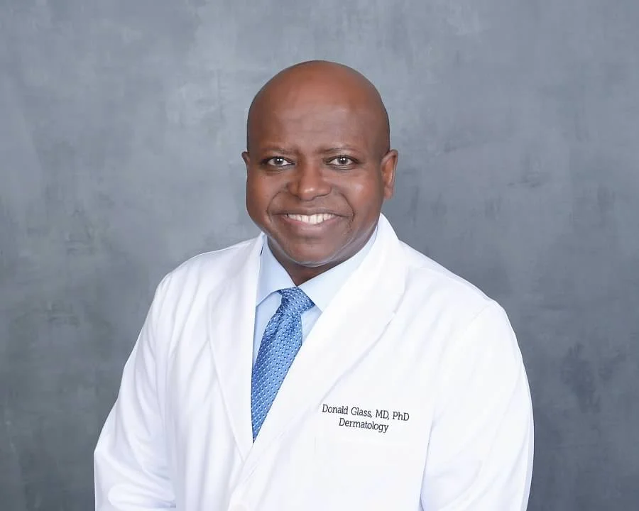 A smiling man in a white medical coat and a blue tie standing against a gray background.