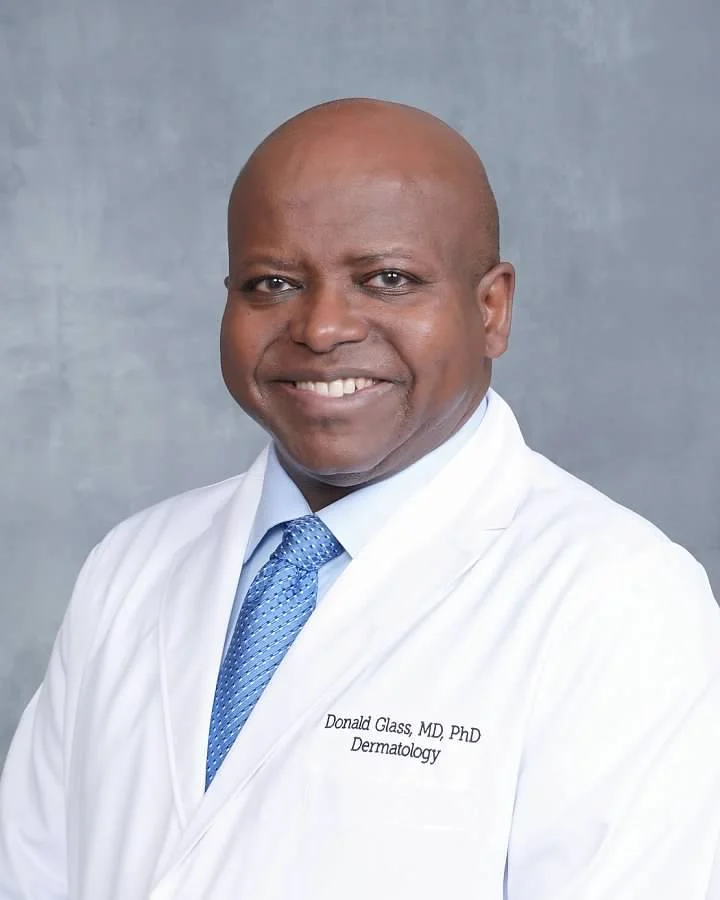 Portrait of a smiling African American man in a white medical coat with blue tie, against gray background.