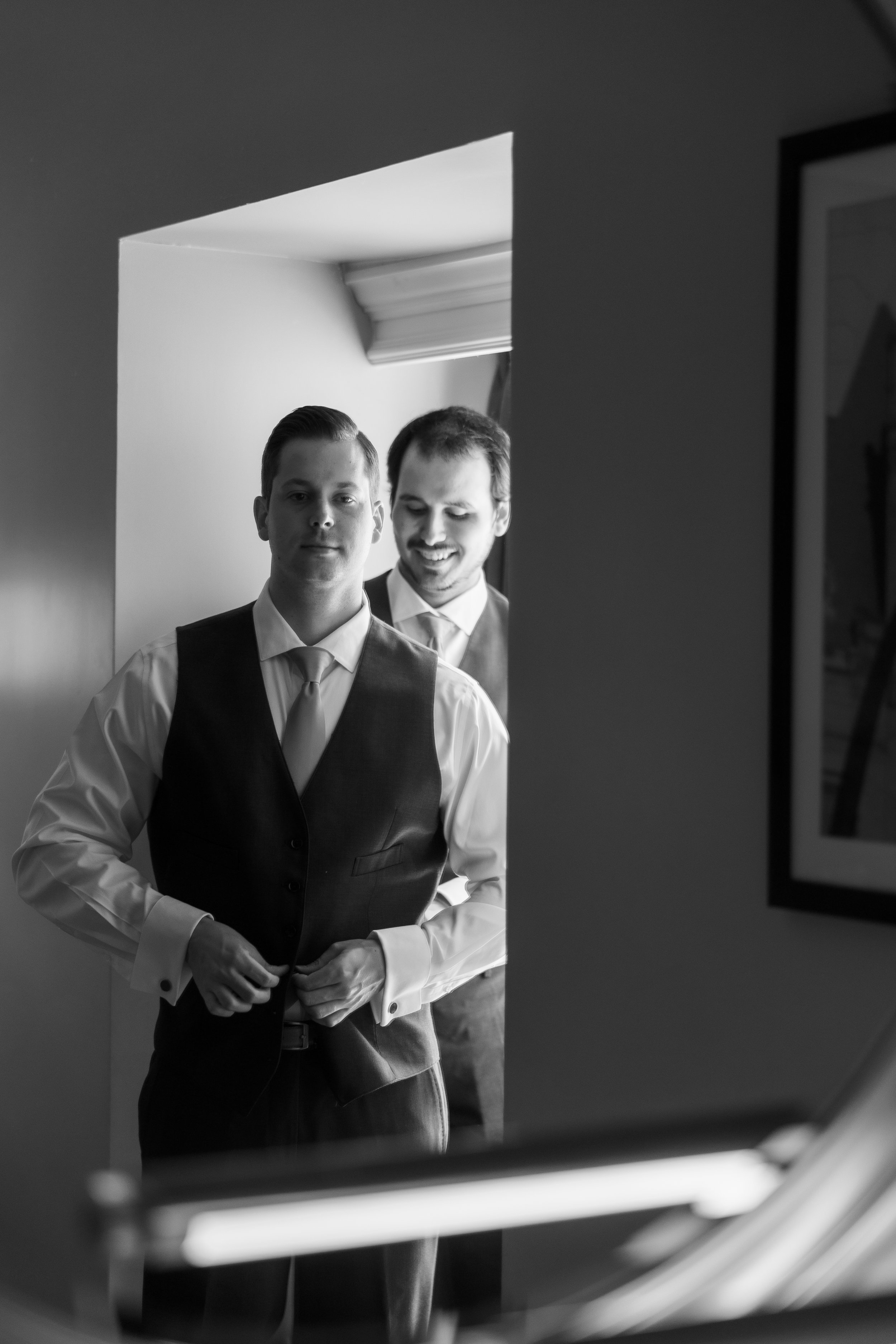 Two young men in formal wear, standing in front of a mirror, adjusting their jackets, smiling and looking at each other, reflected in the mirror.