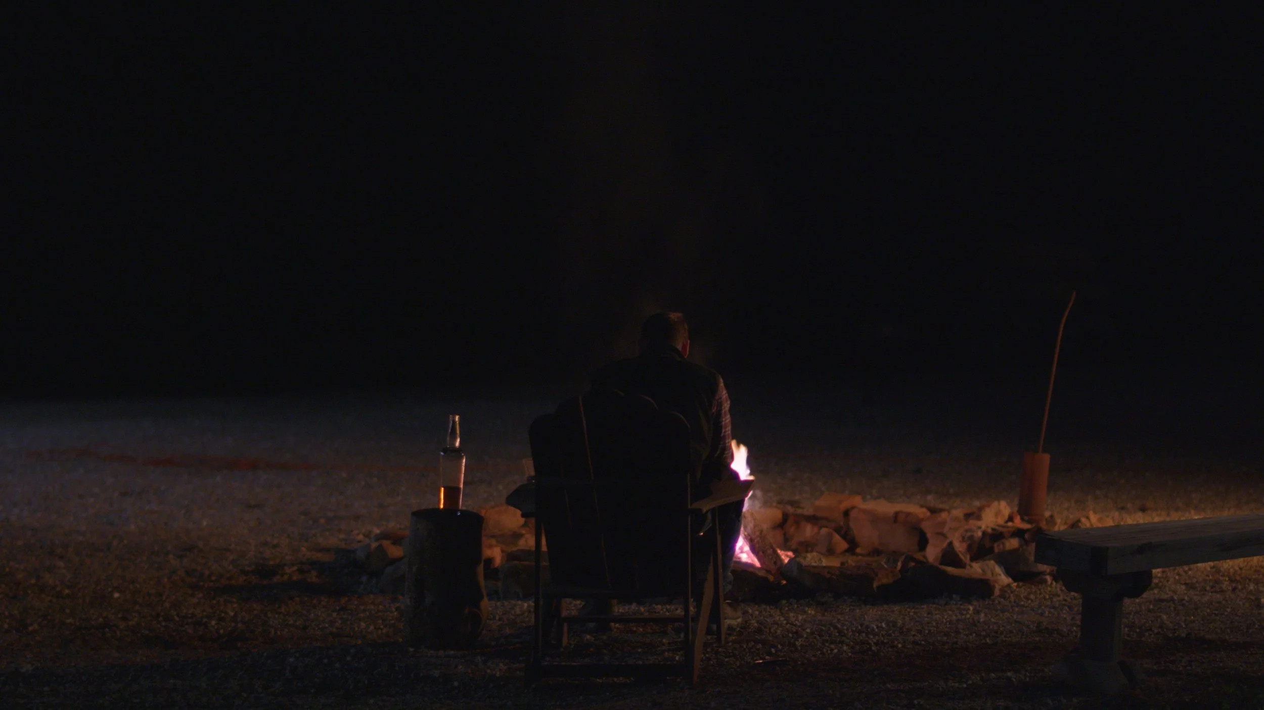Person sitting on a chair by the beach at night, near a fire pit with a drink bottle nearby.