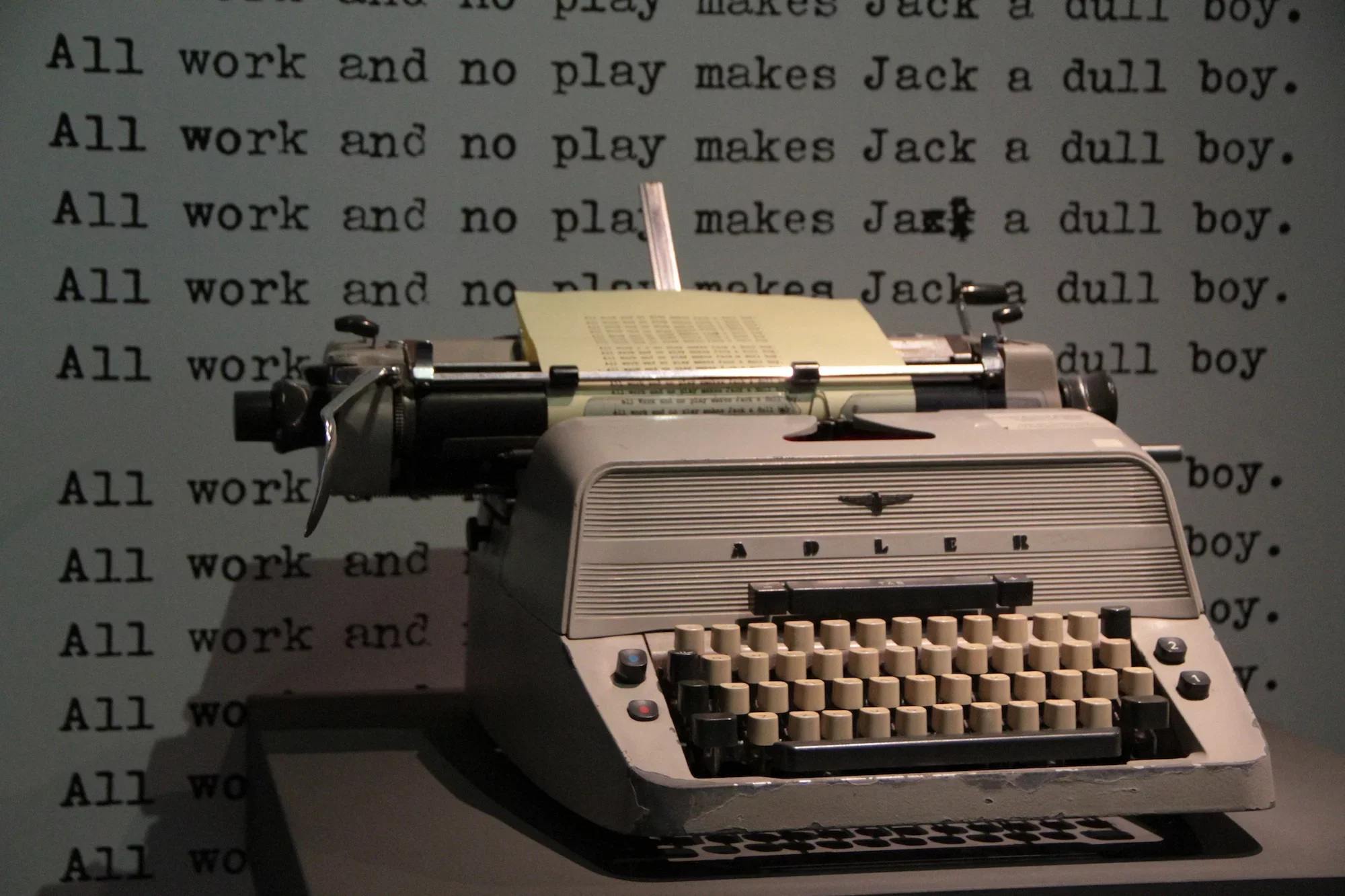 A vintage typewriter with a sheet of paper, set against a background displaying the repeated phrase 'All work and no play makes Jack a dull boy' in black text.