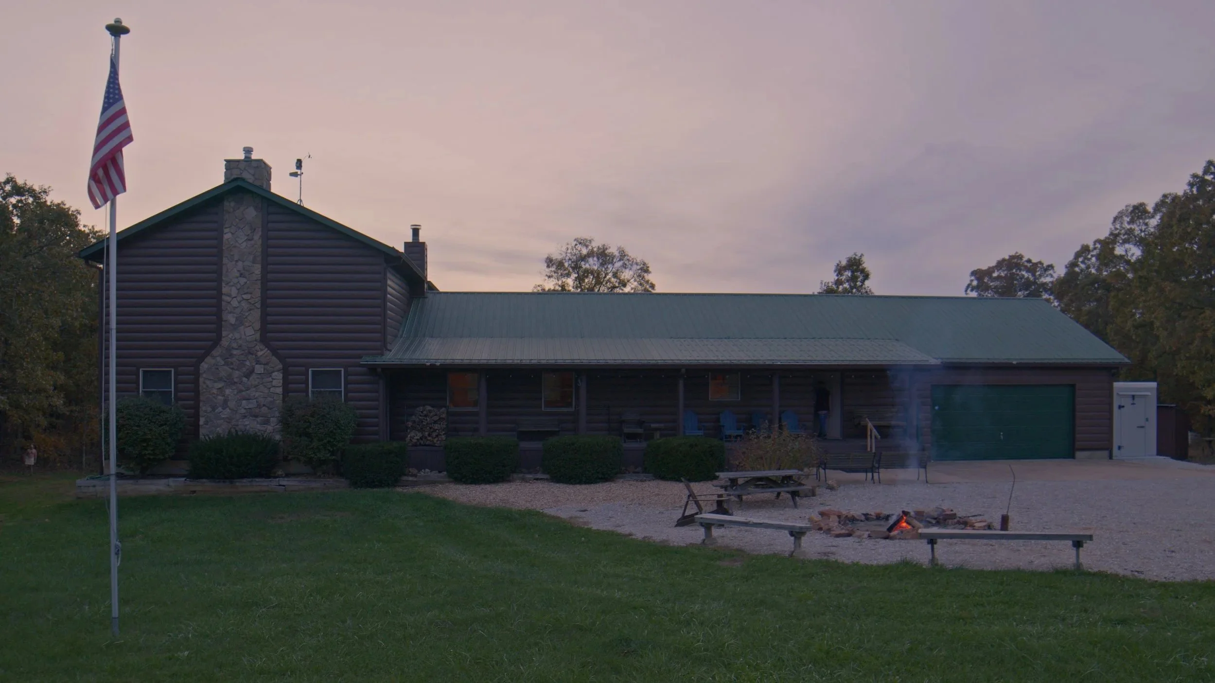 A rustic house with a stone chimney, green metal roof, and wooden siding, surrounded by trees and bushes, with a flagpole flying the American flag in the front yard, and a campfire setup with benches outside.