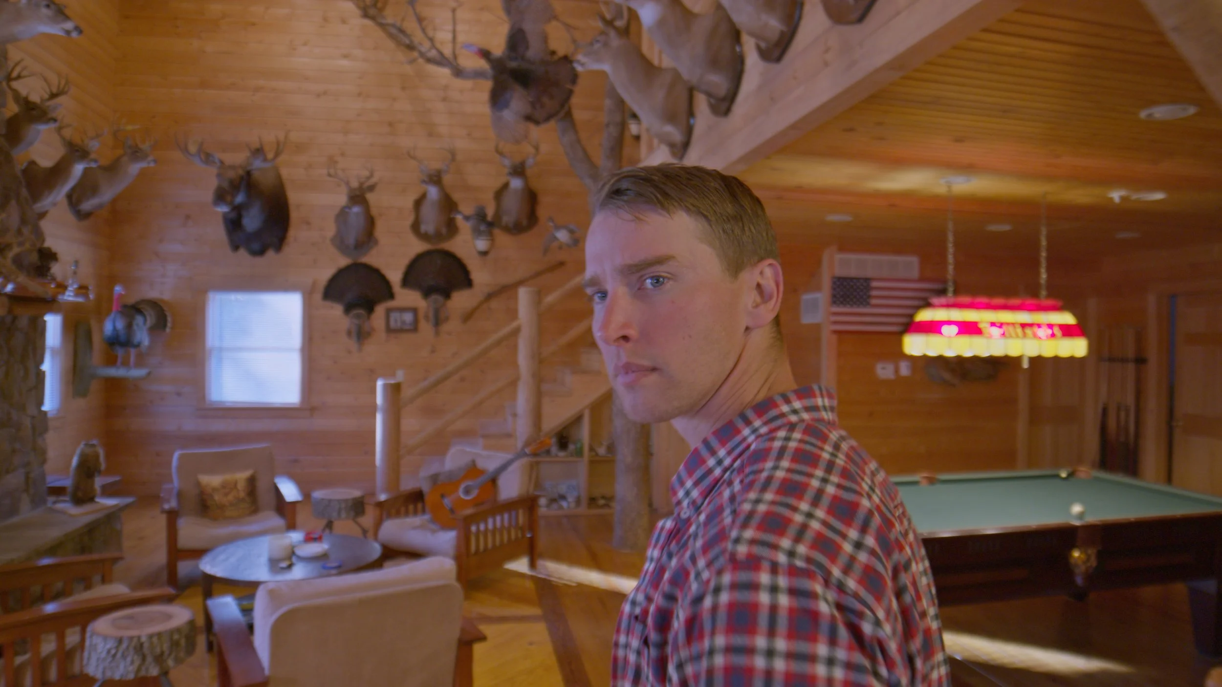 A man with short blond hair and a plaid shirt looking back in a wood-paneled cabin decorated with mounted deer heads, antlers, fans, and American flag wall art, with a pool table and cozy seating area visible in the background.