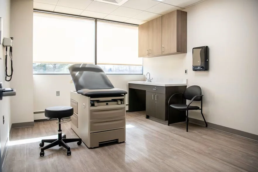 Hospital examination room with an exam table, a stool, a sink, cabinets, a wall-mounted monitor, and a chair, illuminated by natural light from large windows.