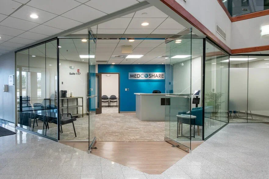 Indoor medical office reception area with glass walls, a reception desk, chairs, and a blue wall with a sign that reads 'MED COST SHARE'.