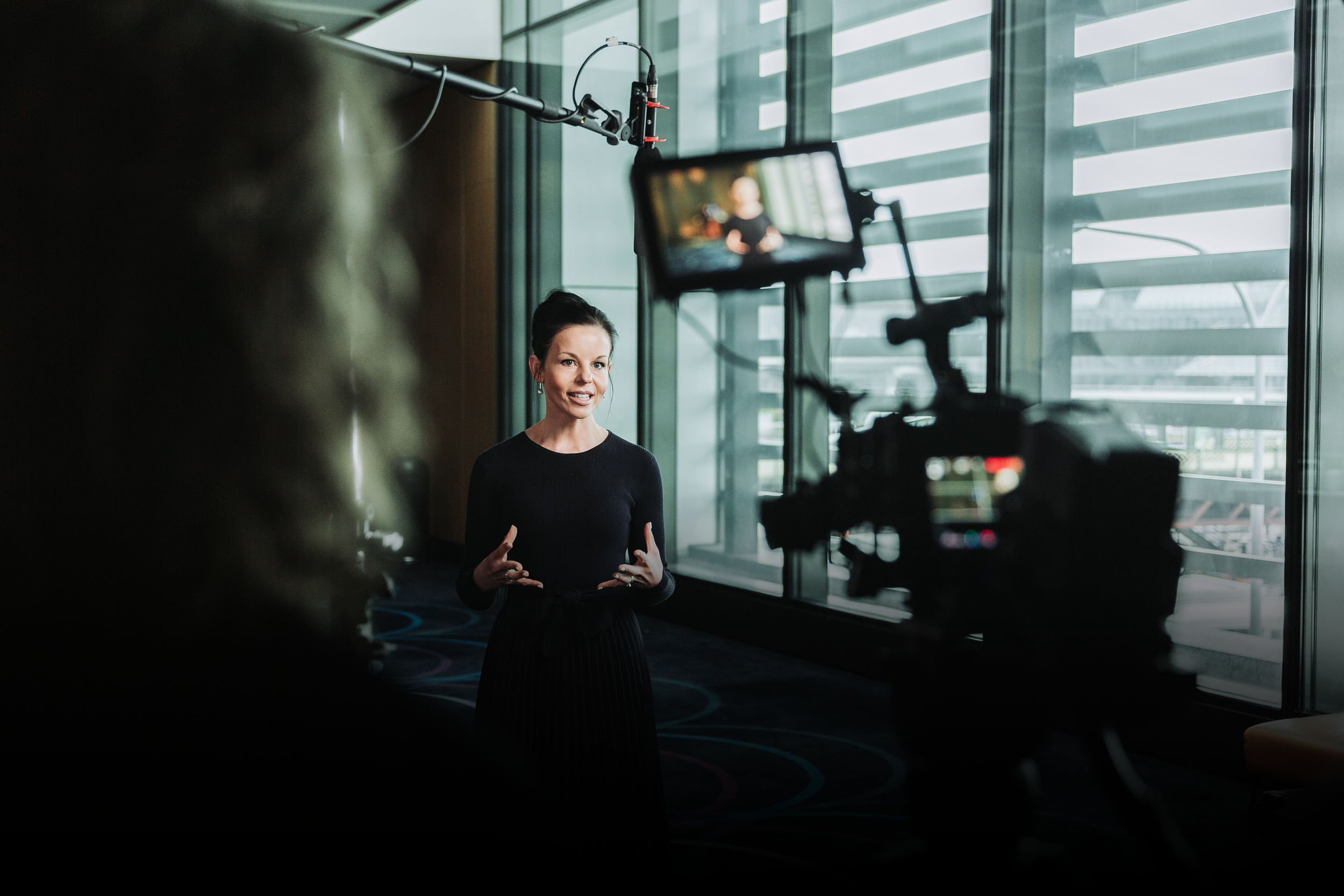 A woman in a black outfit is speaking to a camera in a modern indoor setting with large glass windows.