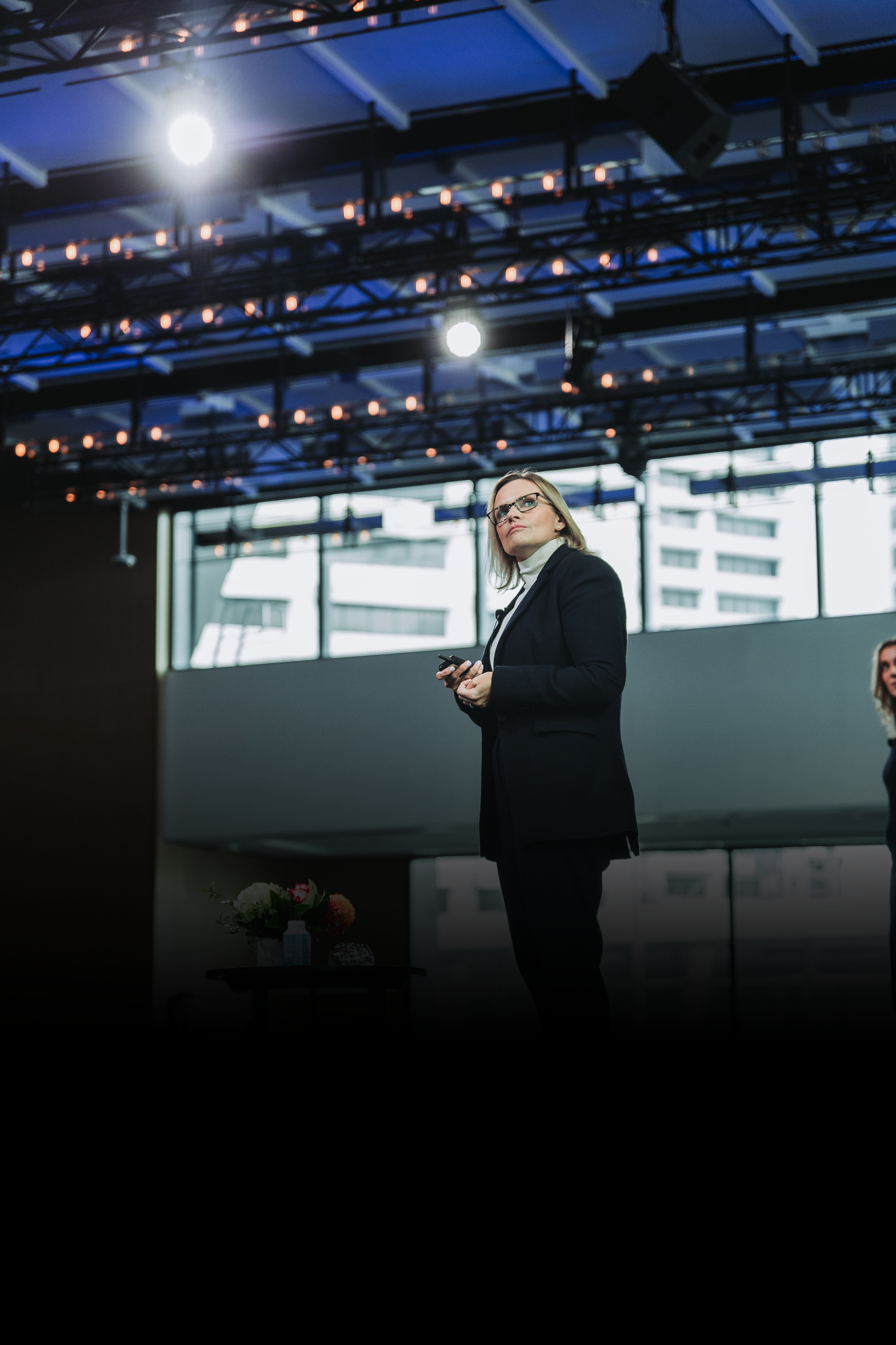 A woman in a black blazer and glasses standing on stage during a presentation or conference, holding a remote control, with a large window and city buildings in the background.