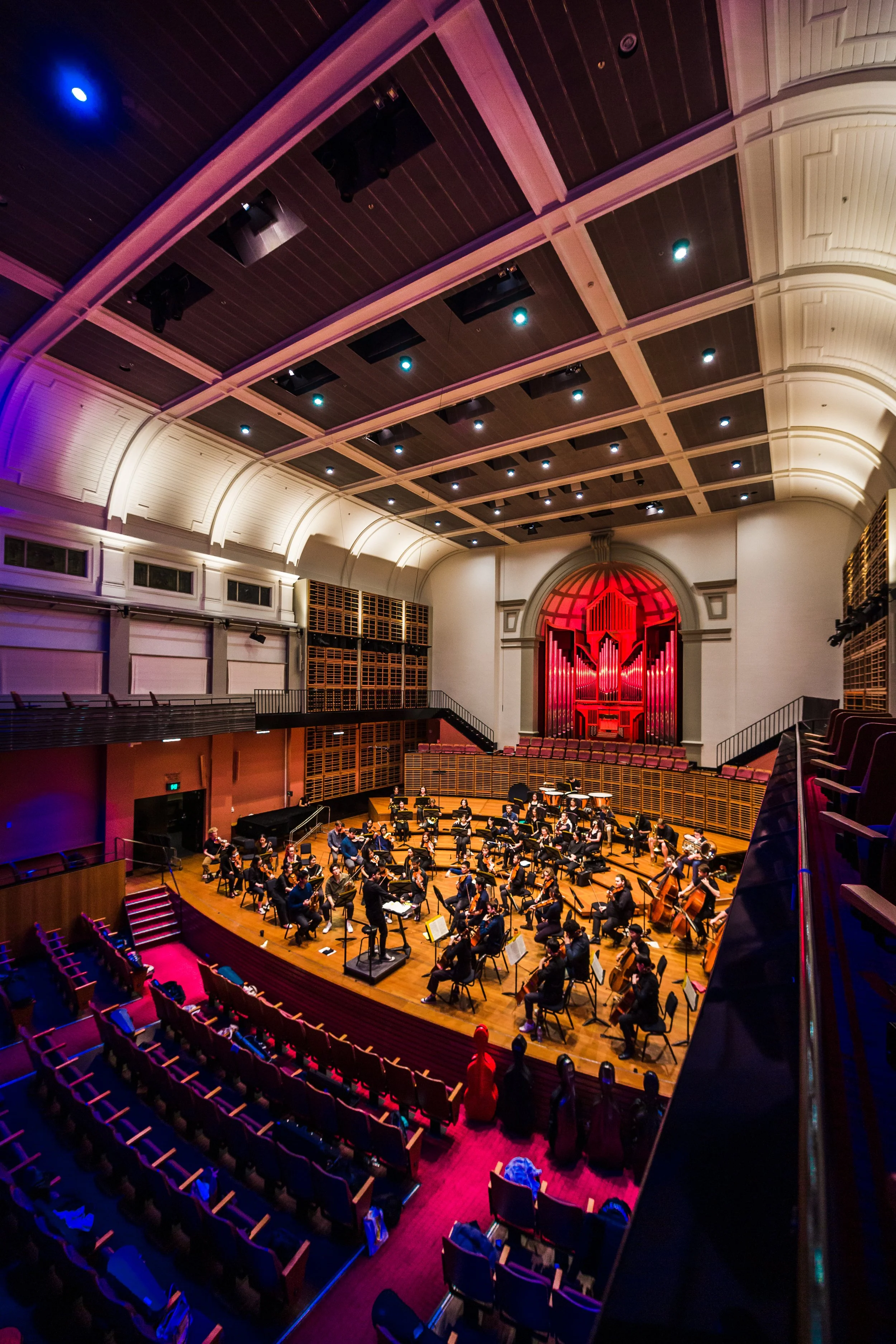 Orchestra performing on stage in a concert hall with seating and organ pipes in the background, illuminated with colorful lighting.