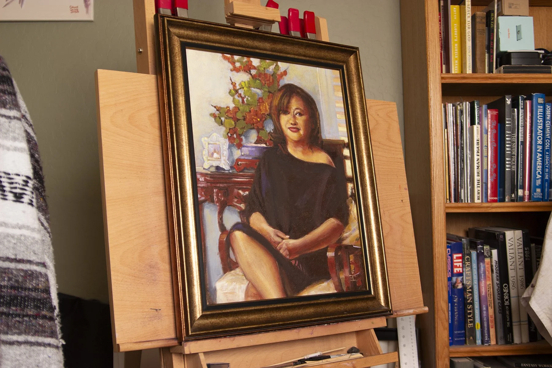 A framed painting of a woman sitting in a chair with one leg crossed over the other. She has shoulder-length dark hair and is wearing a black dress. The background depicts a room with a wooden table, a vase with flowers, and a window with blinds. The