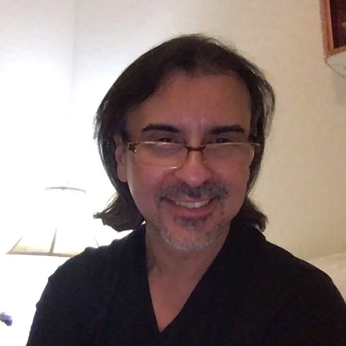 A smiling man with medium-length dark hair, wearing glasses and a black shirt, taking a selfie indoors with a lamp and a wall in the background.