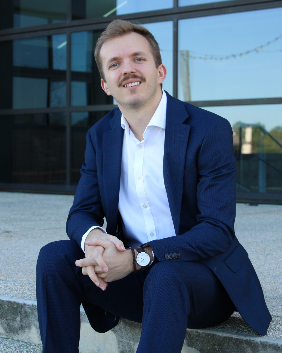 A man in a navy blue suit and white shirt sitting outdoors on a concrete step in front of a modern glass building. He has light brown hair, a mustache, and is smiling. He is wearing a wristwatch and has his hands clasped together.