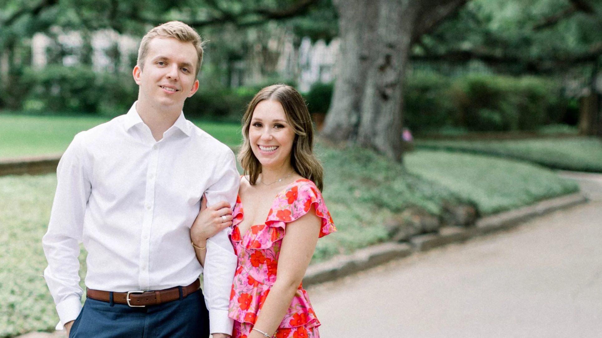 A smiling couple stands outdoors on a sidewalk in a park.