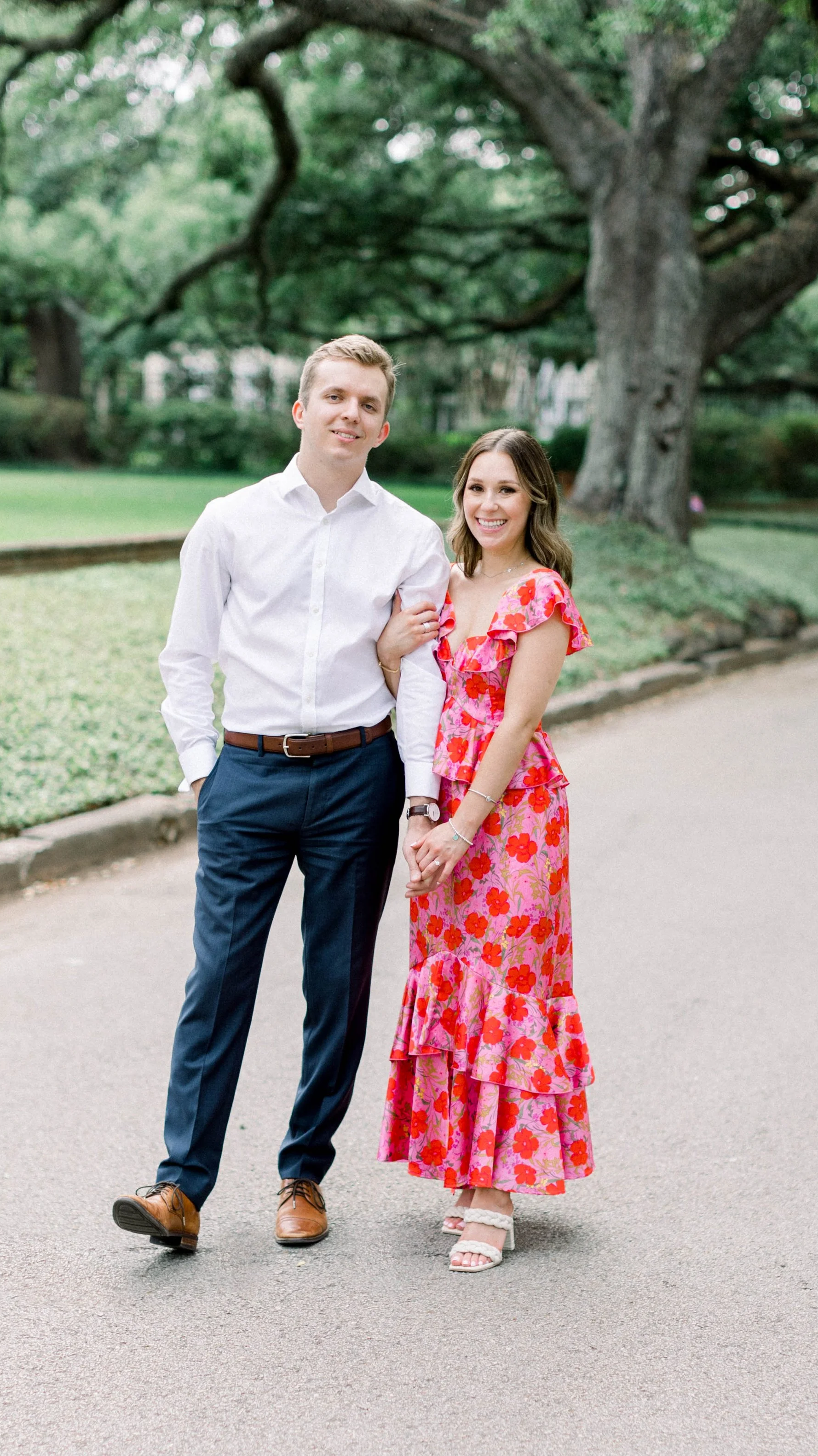 A young man in a white shirt and dark pants holding hands with a young woman in a pink floral dress, standing under a large tree in a park.