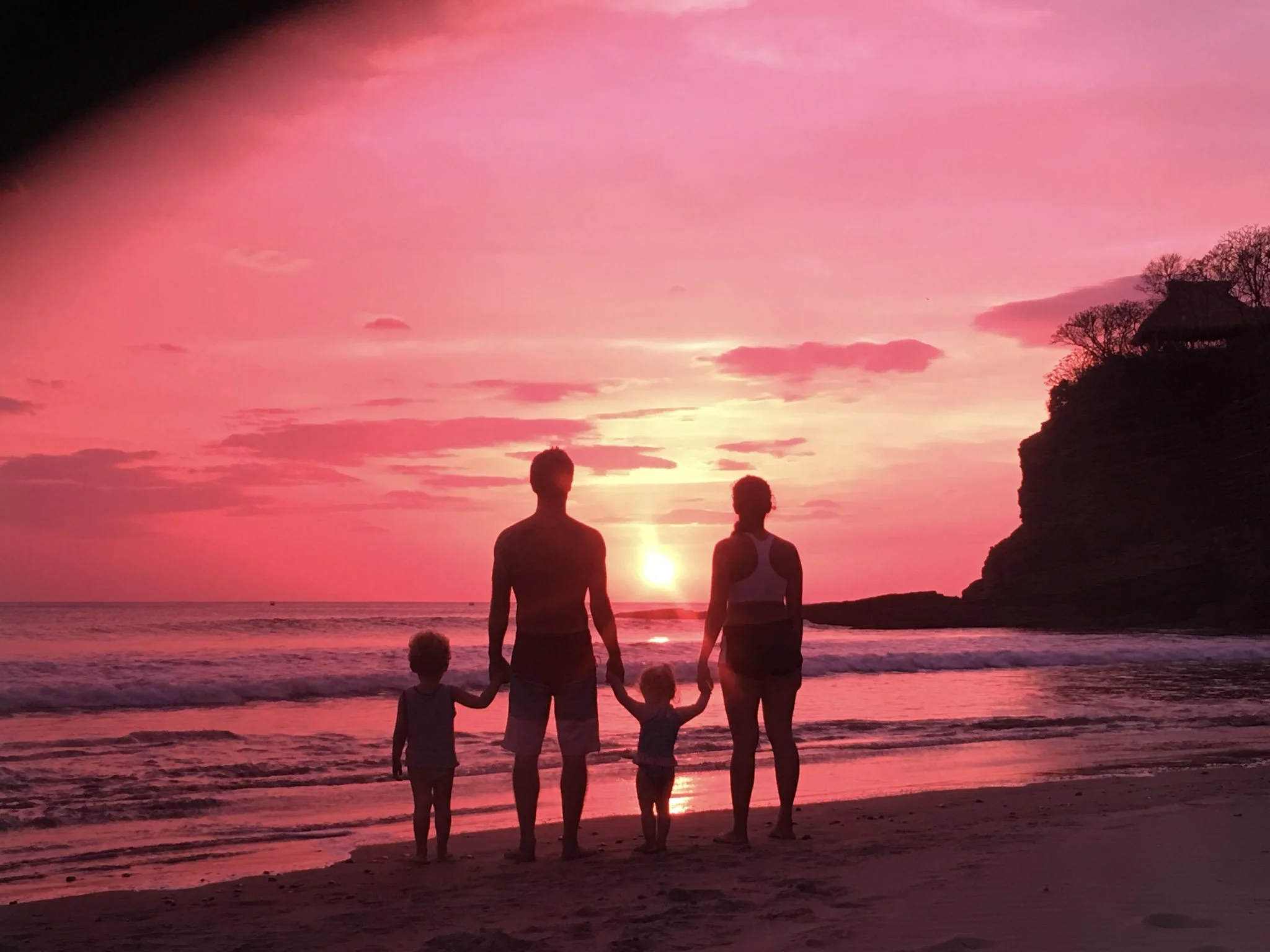 A family of four holding hands on Escameca grande beach at sunset, silhouetted against a pink and orange sky with the ocean and cliffs in the background.