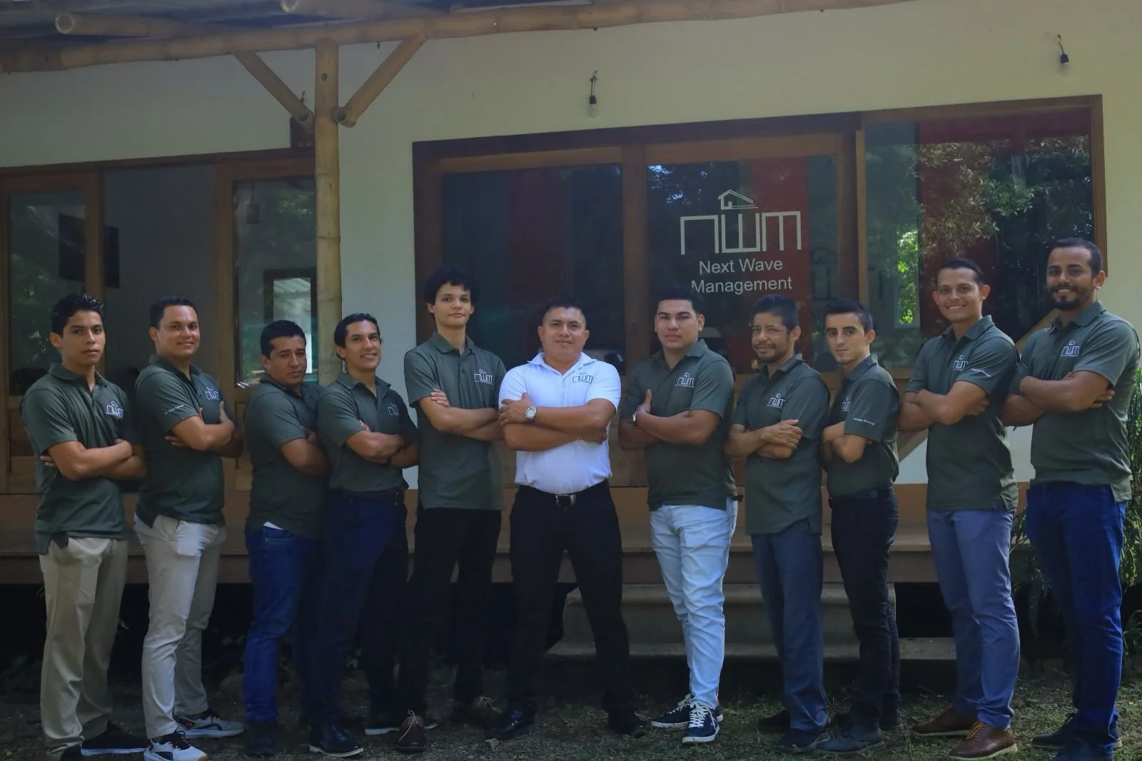 Group of eleven men standing outdoors in front of a building with a sign that reads 'Next Wave Management'. They are wearing matching dark green polo shirts with a logo, and the man in the middle is in a white shirt. They are standing with arms crossed, smiling, and facing the camera.