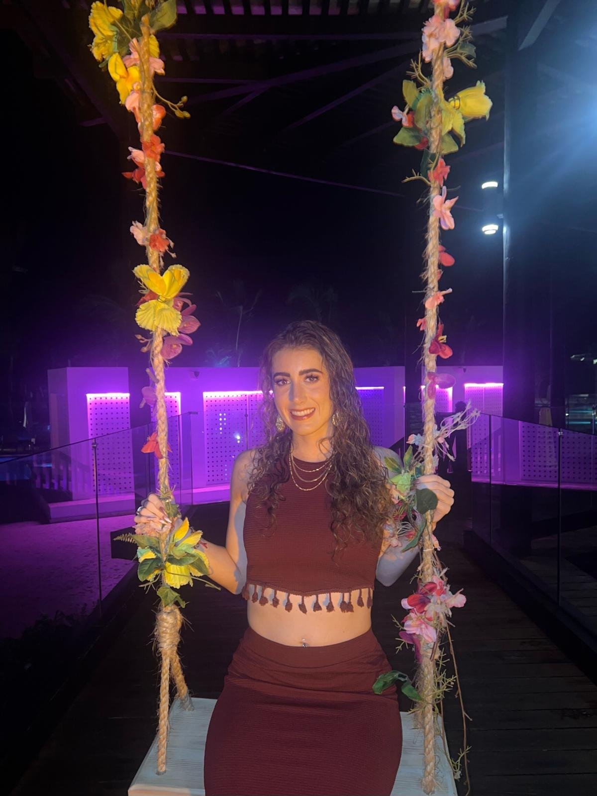 A woman with short blonde hair, wearing a sleeveless orange top and beige pants, is sitting on a bench writing in a notebook. Sunlight streams in through a window, casting shadows on the wall behind her and a piece of abstract art leaning against the wall on her right.