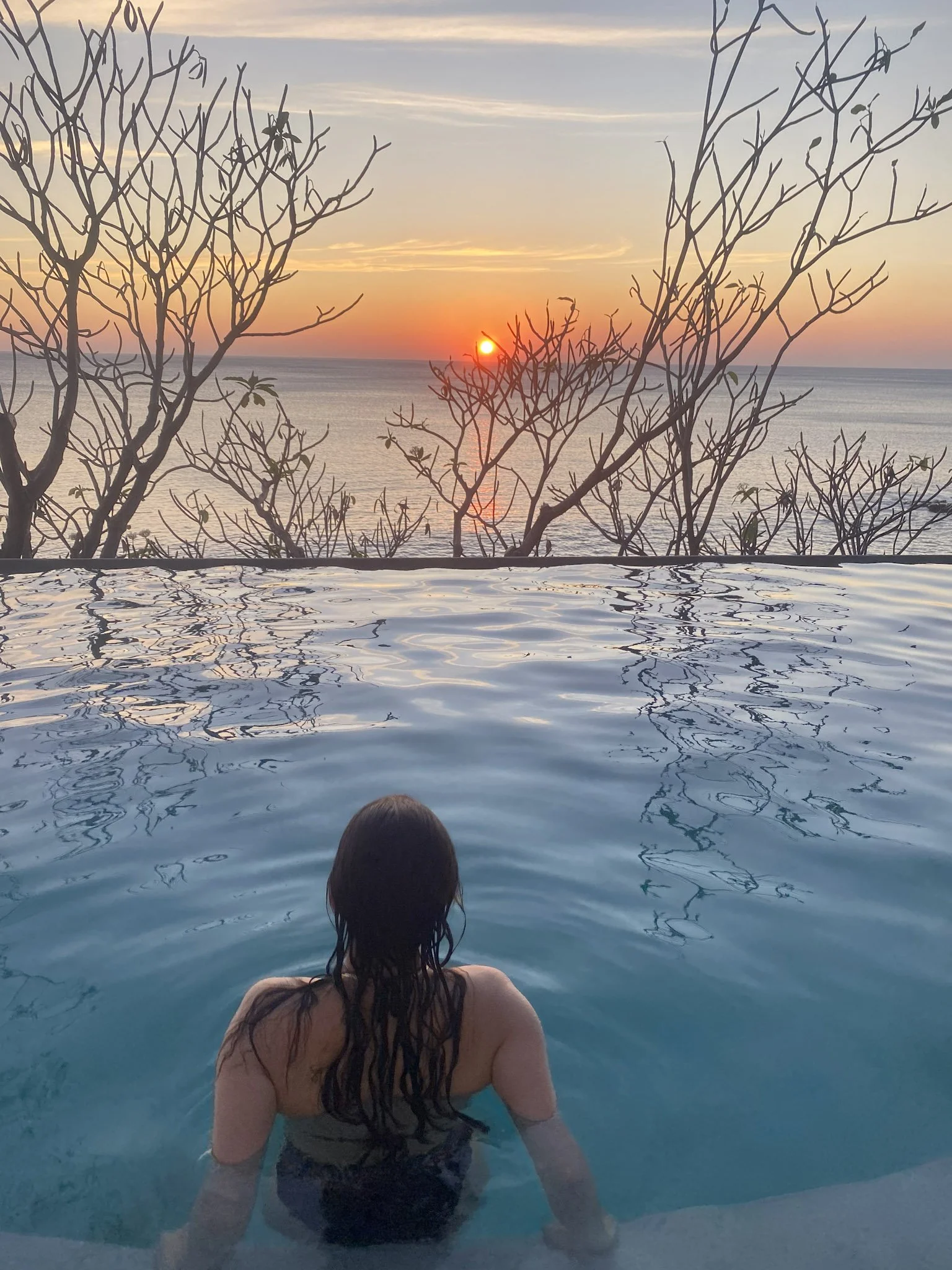 A woman with long dark hair swimming in the La Punta spa infinity pool during sunset, back facing the camera with an ocean and barren trees in the background.