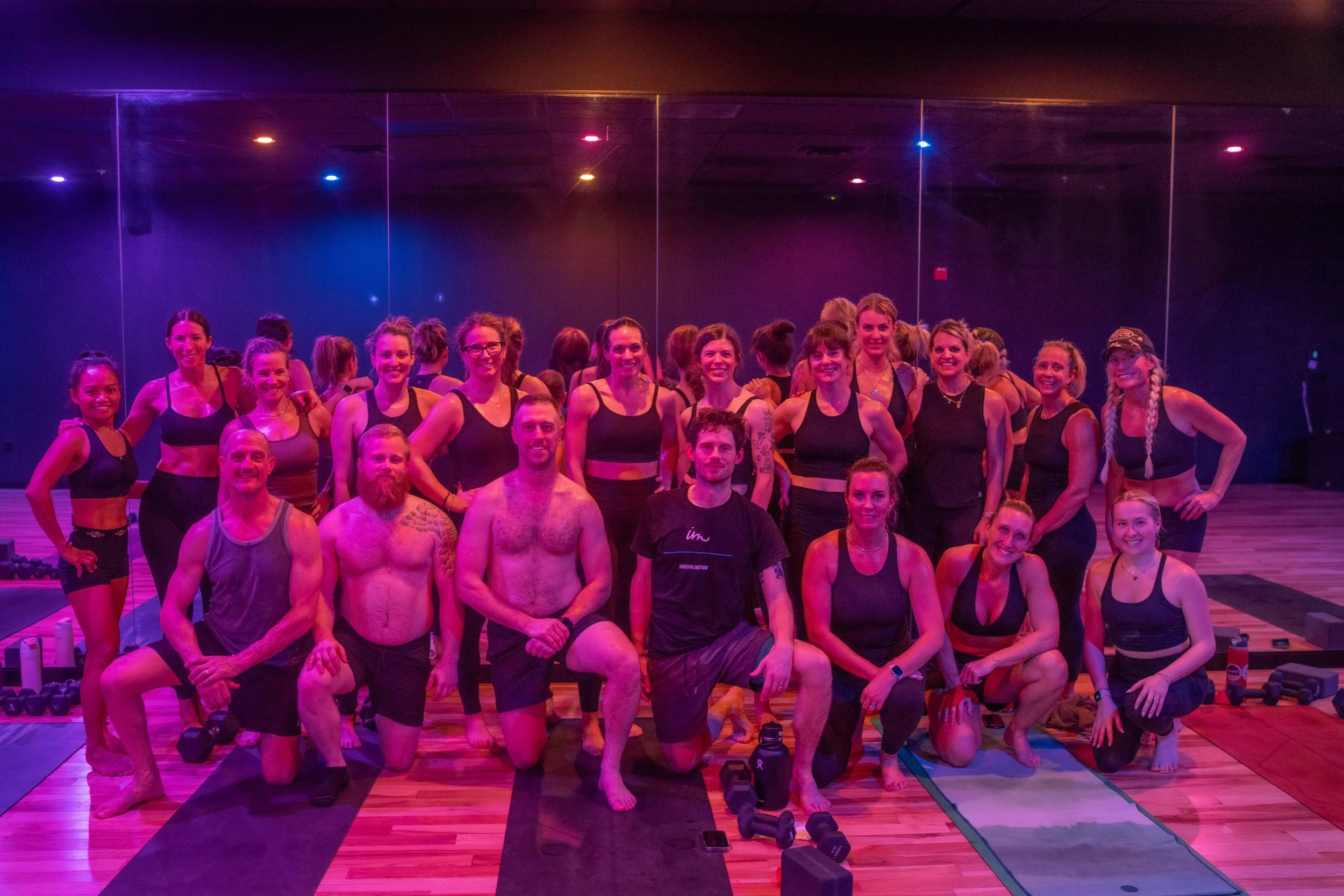 Group of women and men in a fitness class in a dimly lit gym with purple and blue lighting, standing and kneeling on yoga mats, some holding water bottles and workout equipment.