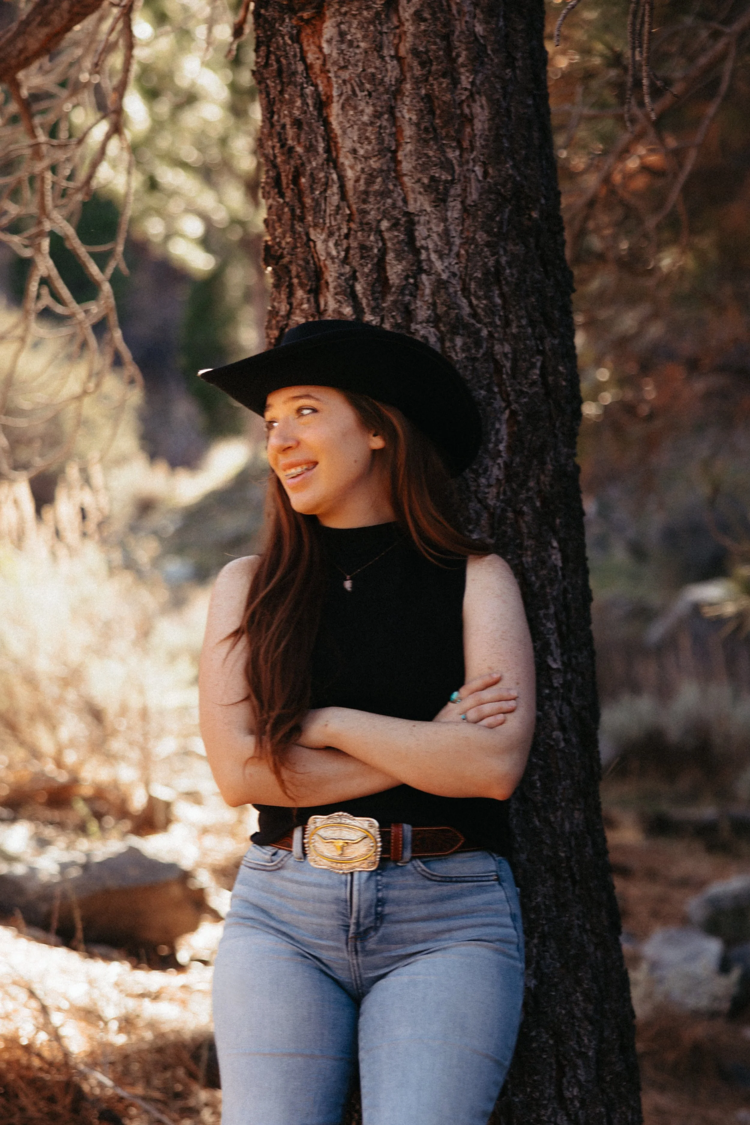 Morgan Wadsworth, candidate for congress wearing a black cowboy hat, black sleeveless top, blue jeans, and a belt with a large buckle, standing outdoors next to a tall tree, with her arms crossed and smiling.