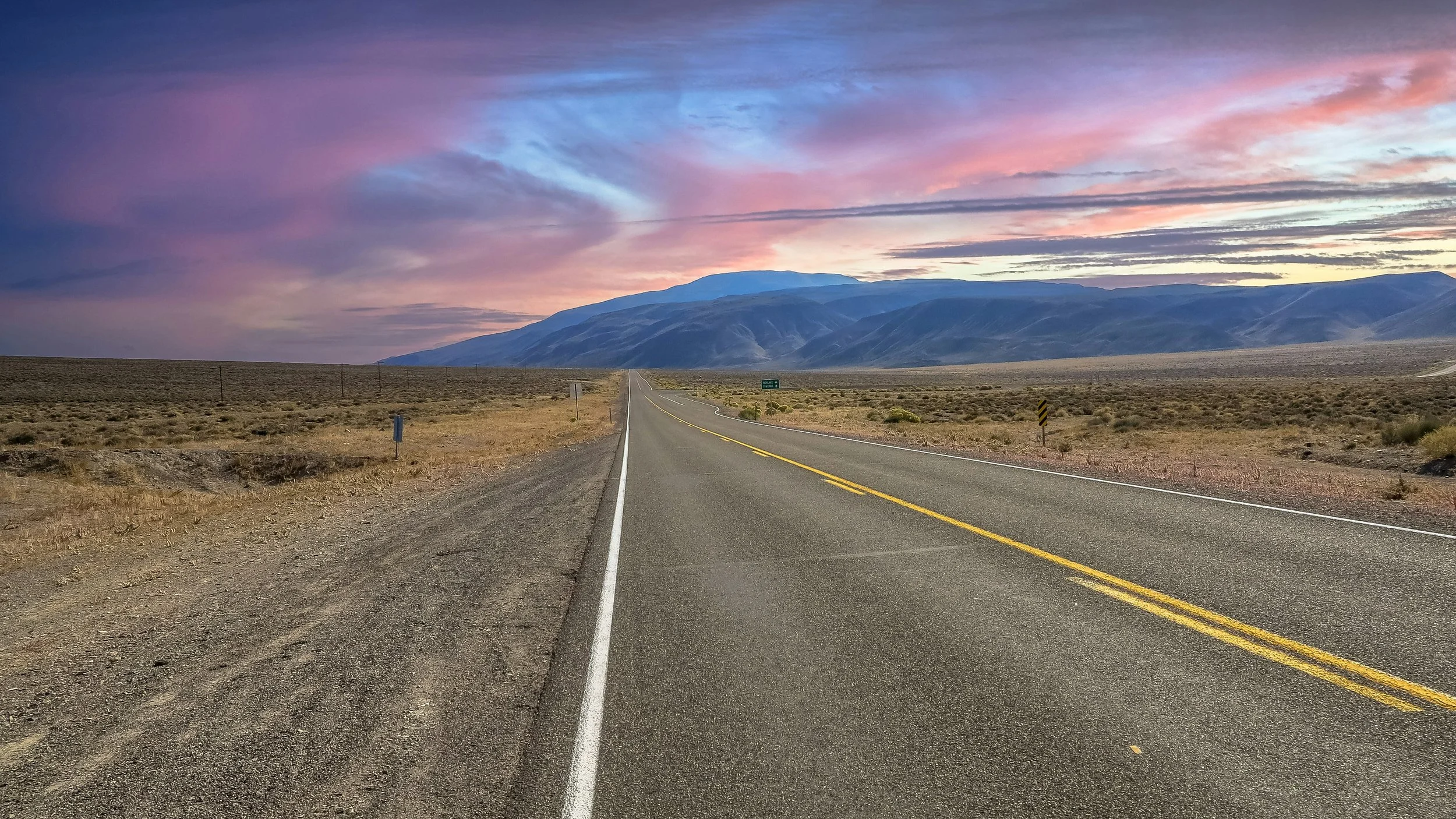 A long empty road running through the Northern Nevada desert landscape with mountains in the distance under a colorful sunset sky.