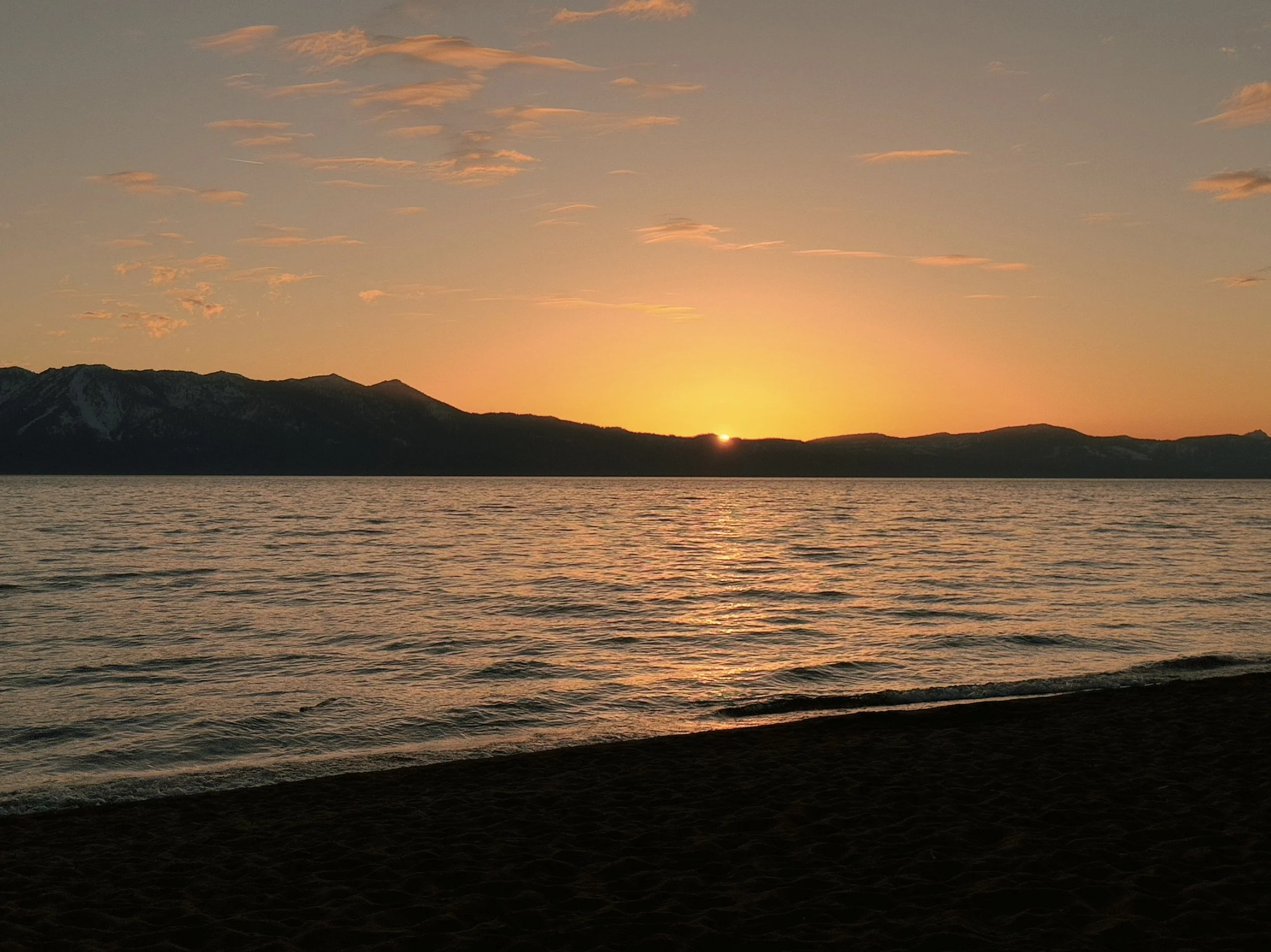 A landscape scene of a sunset over a mountain range, with calm water in the foreground and a partly cloudy sky illuminated by the setting sun.
