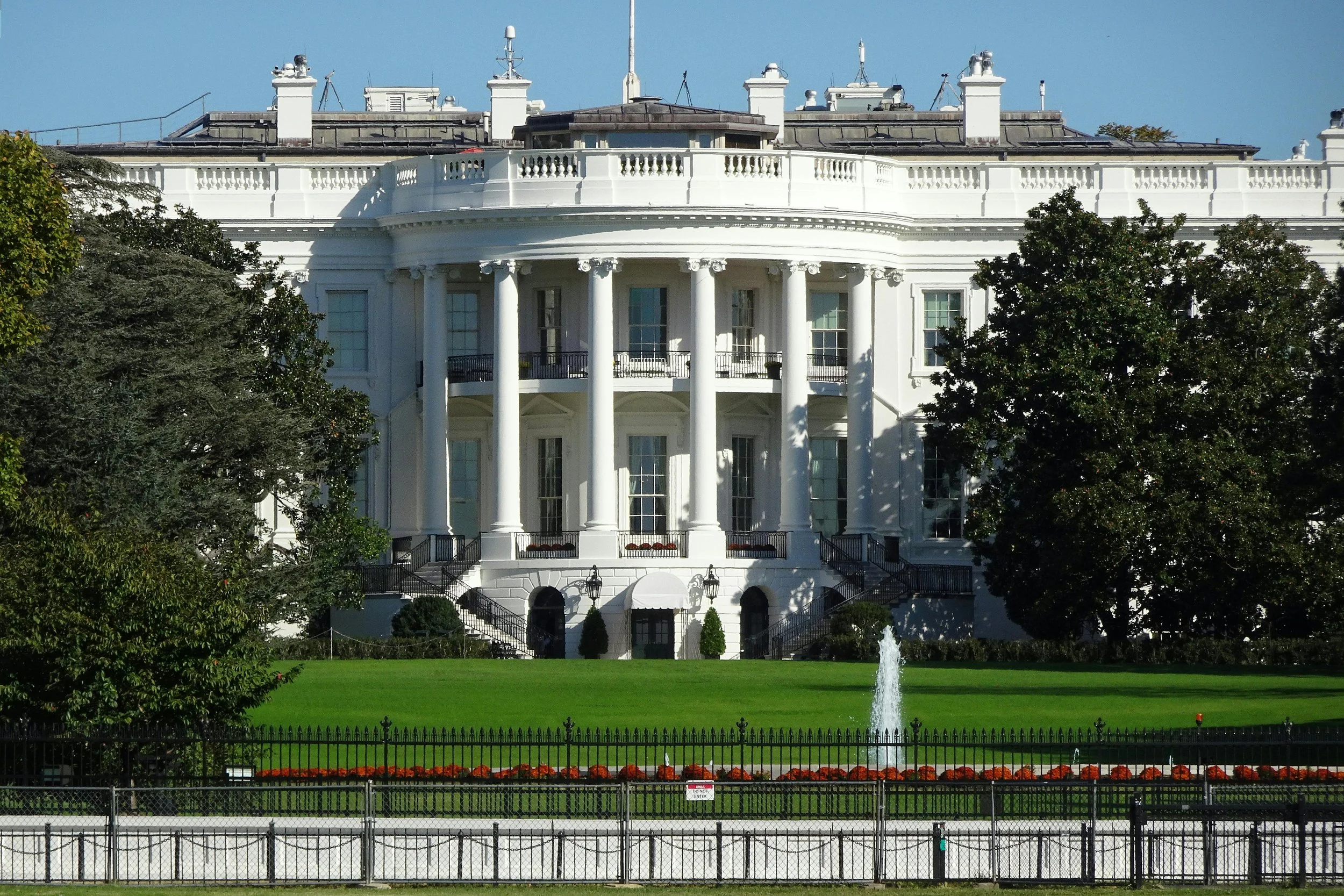 The White House, the official residence and workplace of the President of the United States, located in Washington, D.C., seen here with a fountain in front, surrounded by trees and a black metal fence.