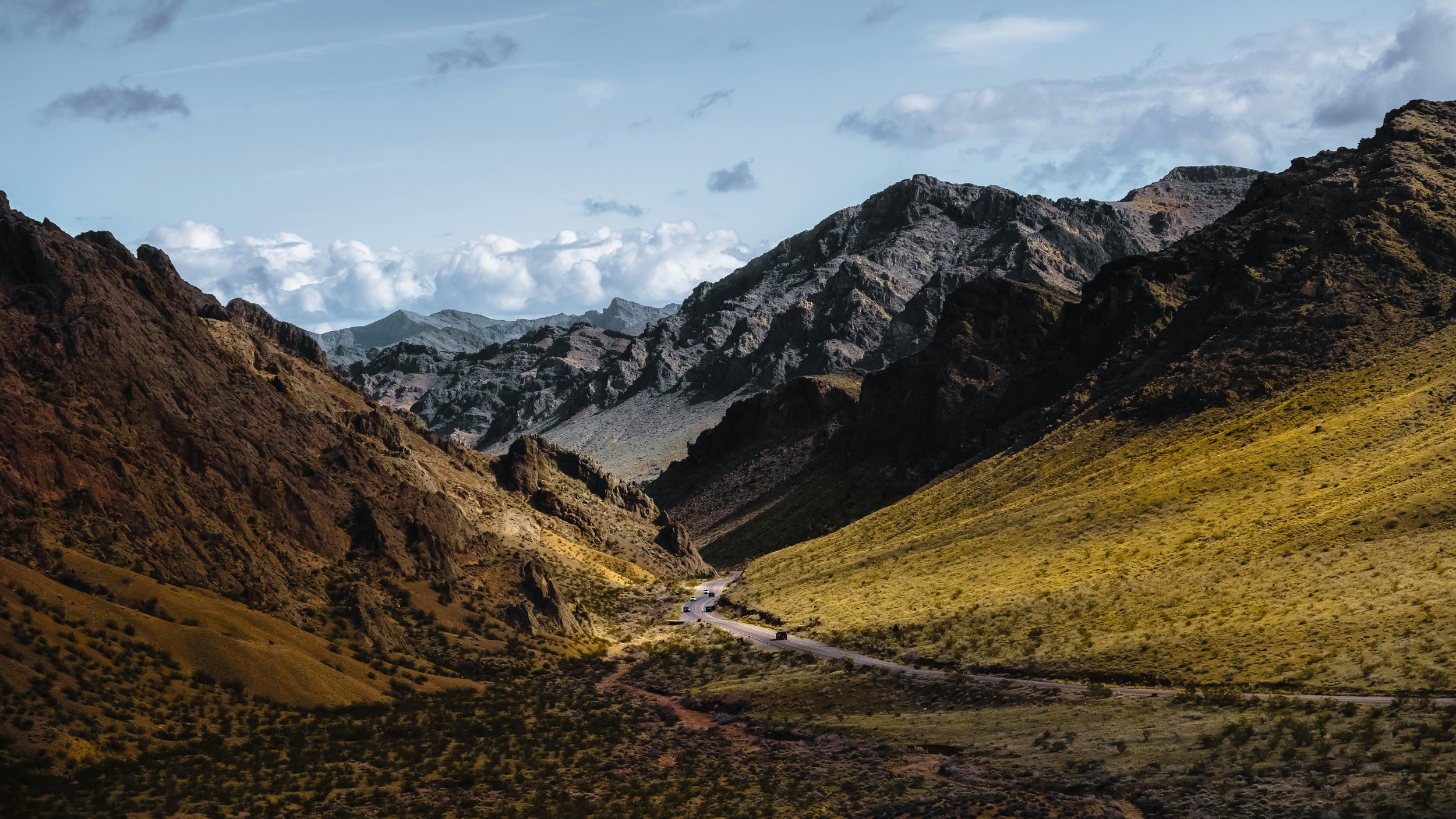 A winding mountain road through a valley in Nevada with rugged, rocky mountains in the background under a partly cloudy sky.