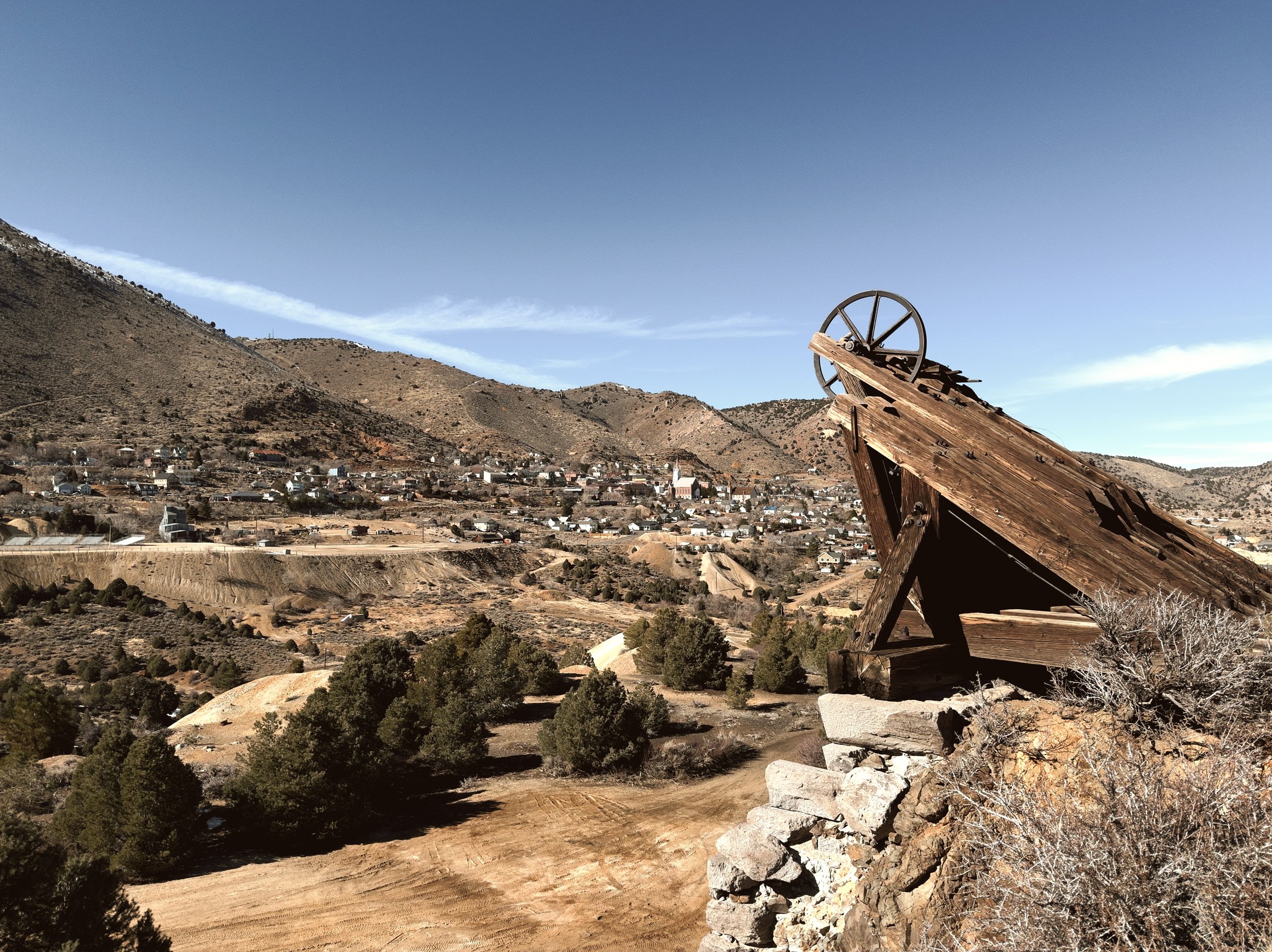 A scenic view of a small town at the base of a hill, with a large, old wooden water wheel in the foreground, under a clear blue sky.
