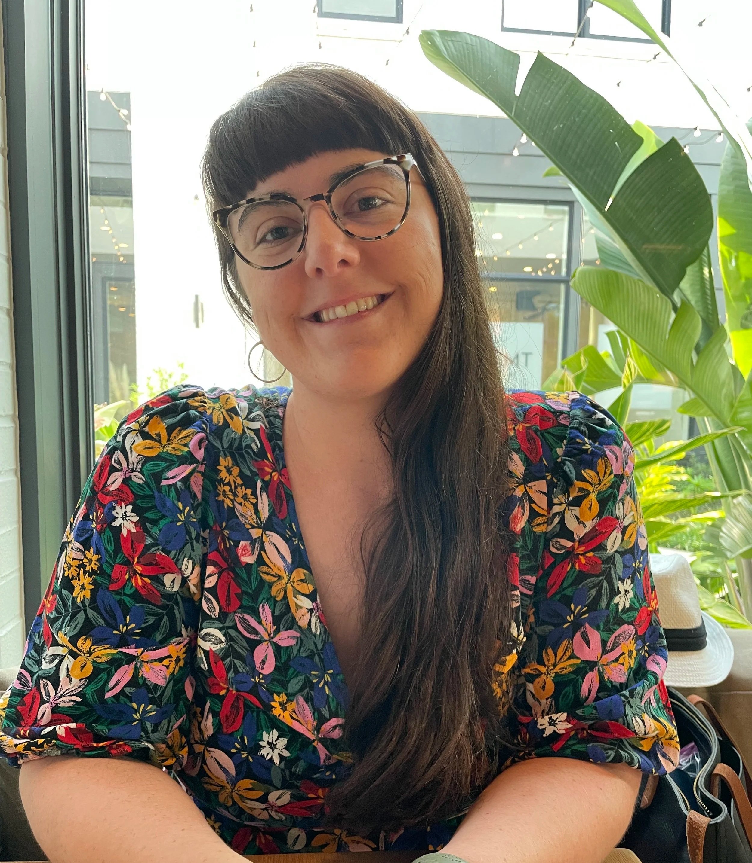 A woman with dark brown hair, bangs, glasses, and earrings, smiling at the camera, wearing a vibrant floral-patterned blouse, sitting indoors near large windows with green plants around.