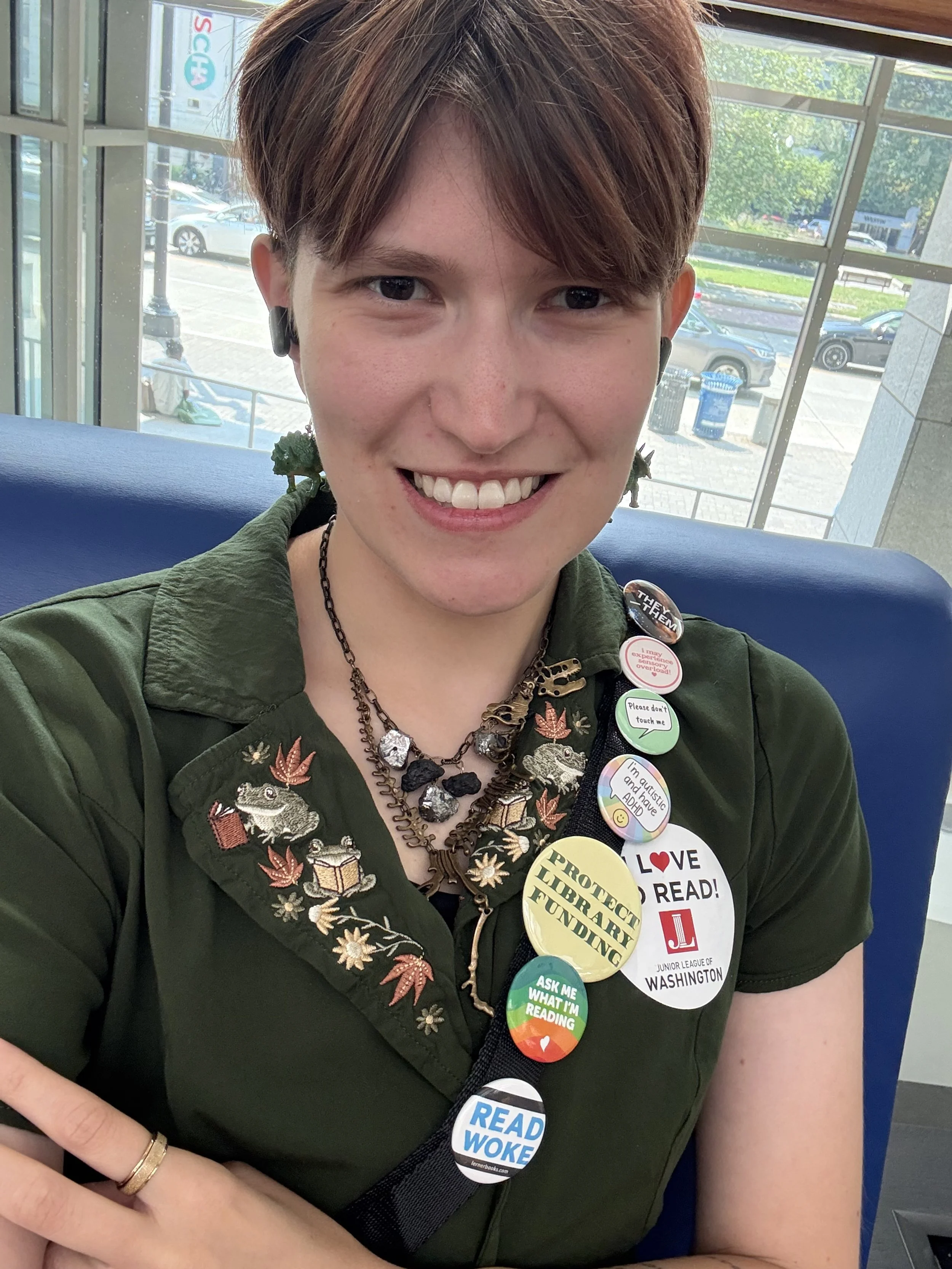 A smiling young woman with short brown hair wearing a green embroidered shirt and multiple colorful buttons and pins, sitting indoors near large windows with a parking lot and trees visible outside.