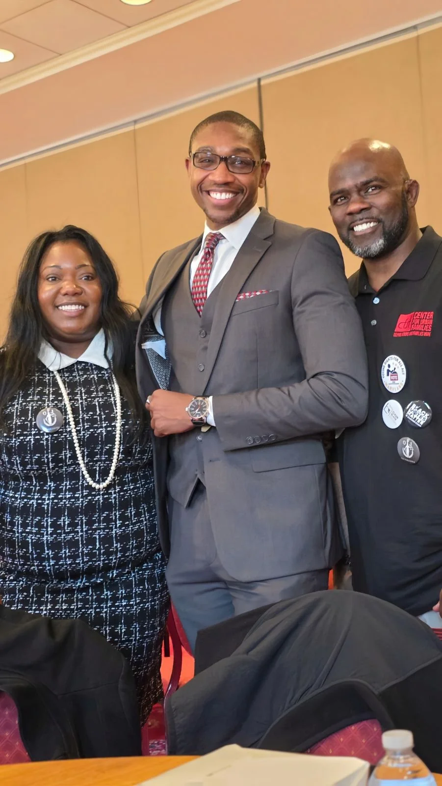 Three smiling people at what appears to be a professional or conference event. The woman on the left is wearing a black and white patterned outfit with a pearl necklace and badge. The man in the middle is wearing a gray suit with a red patterned tie, glasses, and a watch. The man on the right is wearing a black polo shirt with badges and logos, including one that says 'Center for Urban Families'.