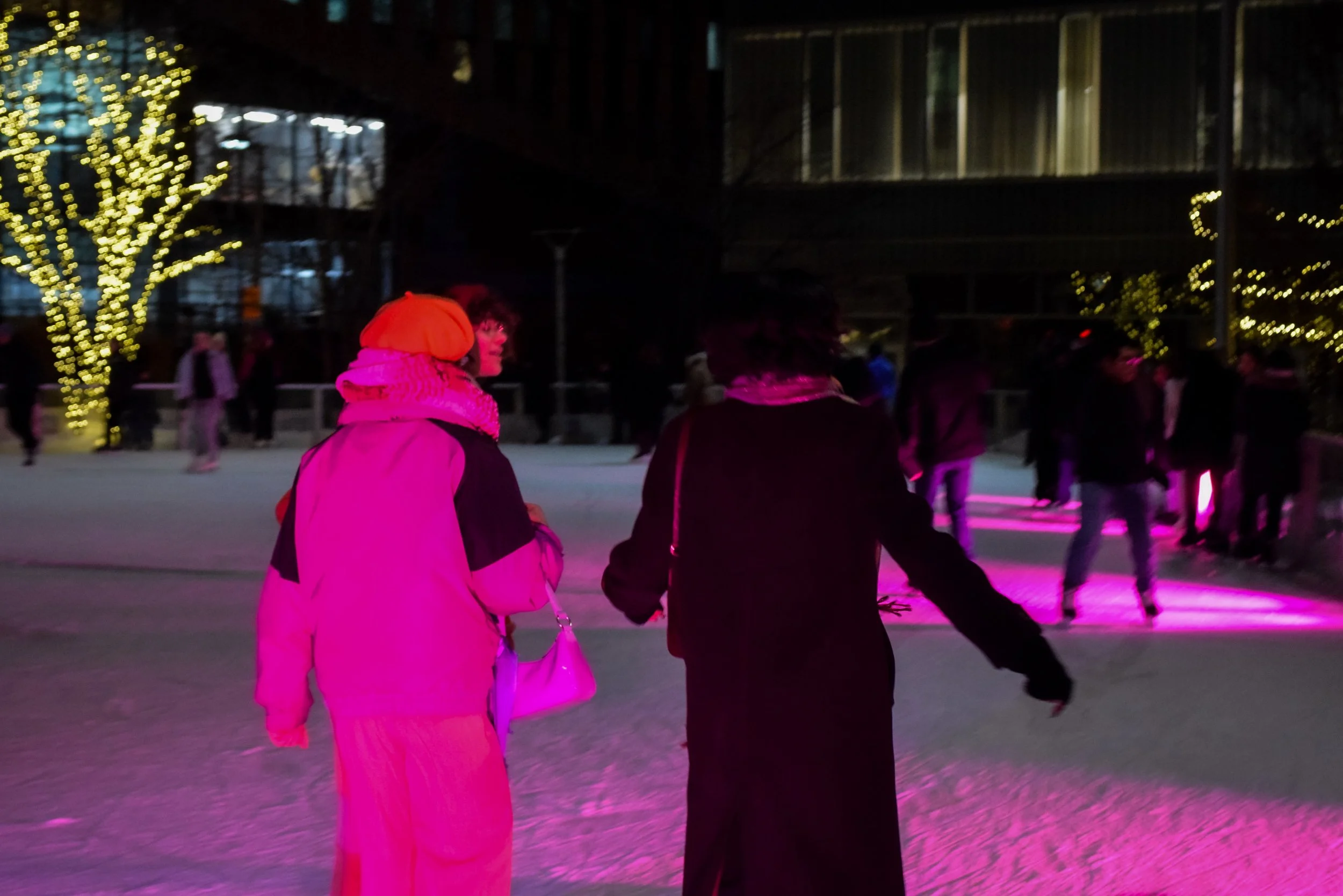 People skating on an ice rink at night with festive lights and modern buildings in the background.
