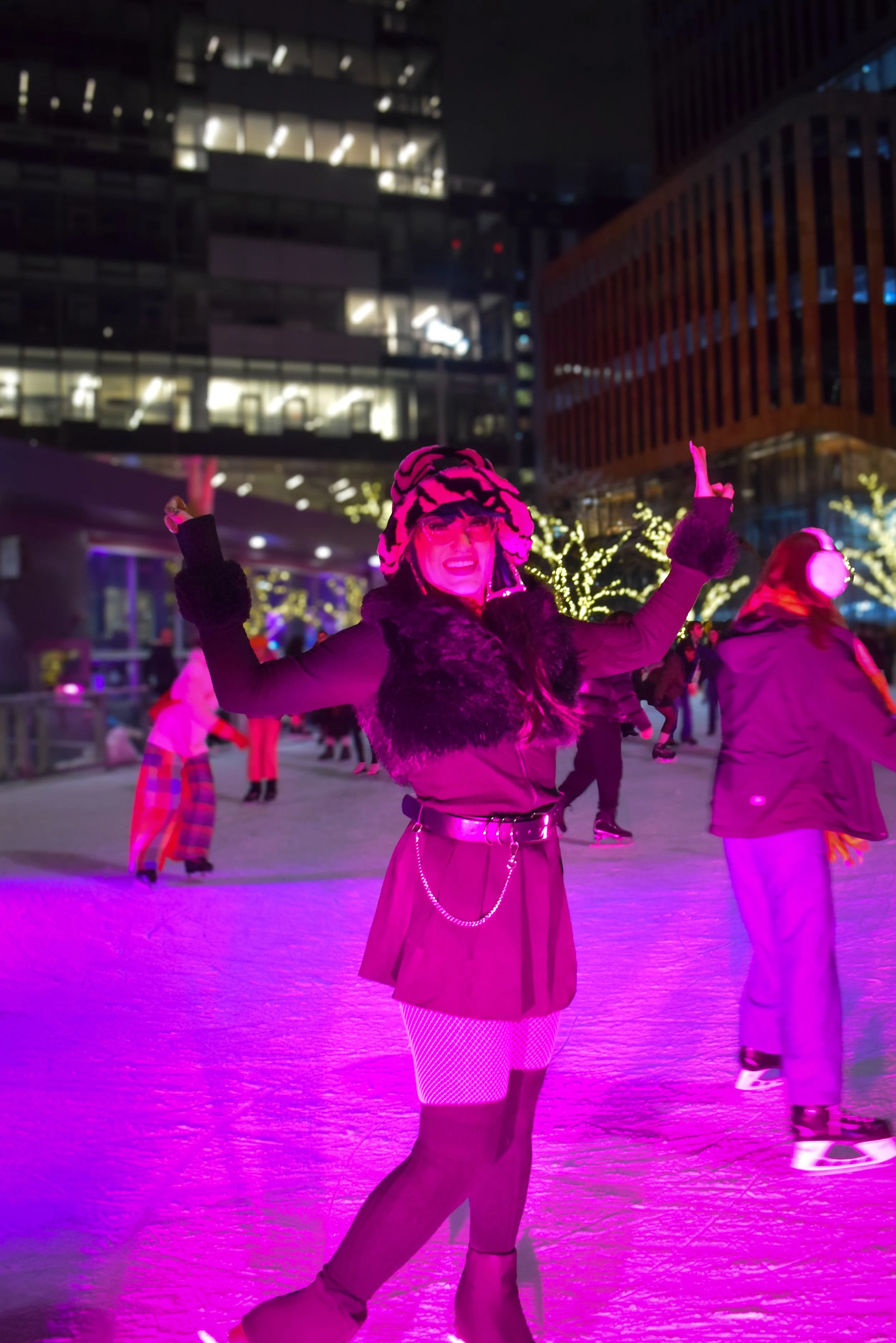 Woman ice skating at night outdoors illuminated by purple and pink lights, dressed in costume with striped tights, fishnet stockings, and a furry hat, smiling, with other skaters and modern buildings in the background.