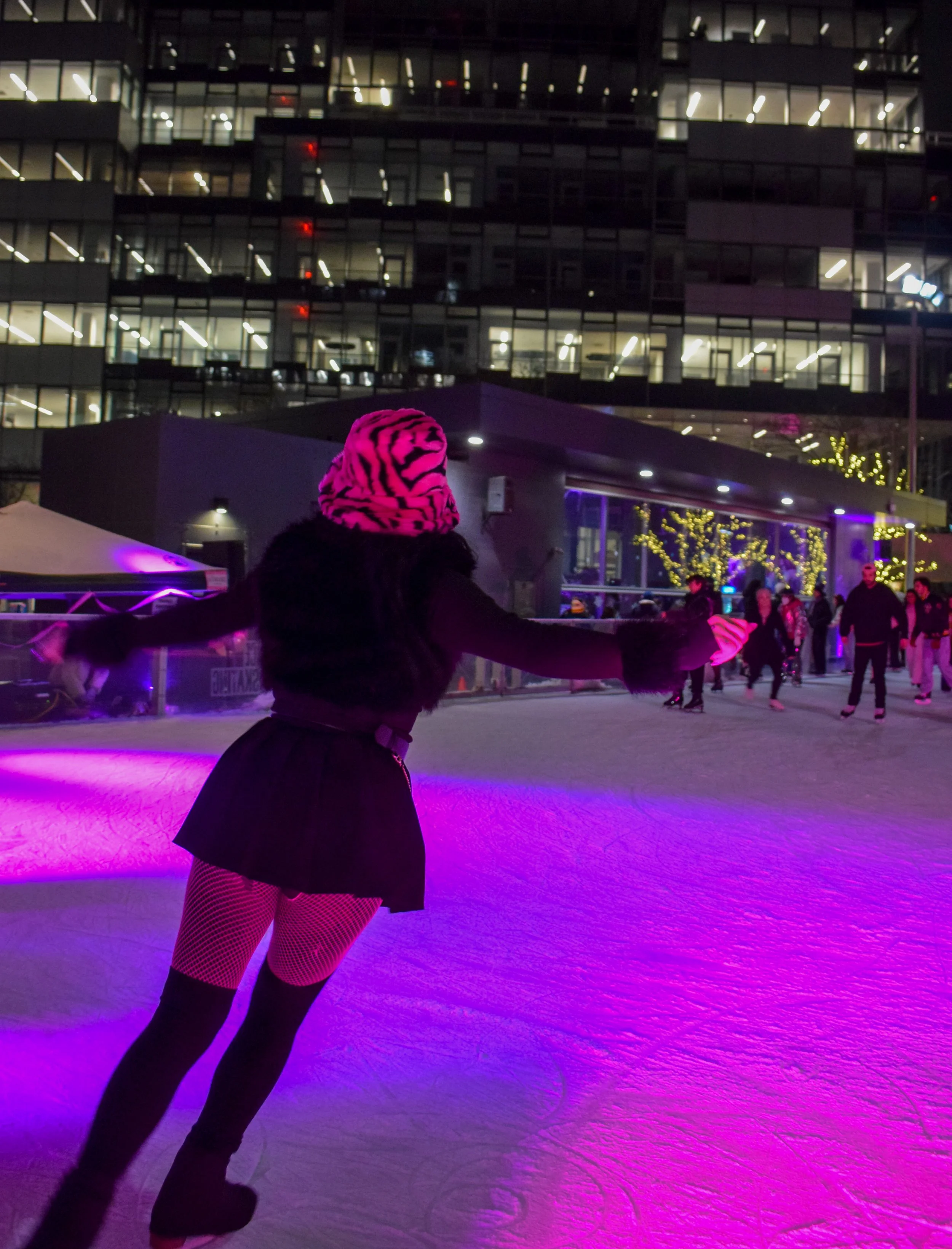 Person ice skating at an indoor rink at night, wearing a pink and black zebra-striped hat and fishnet tights, with a cityscape background and purple lighting on the ice.