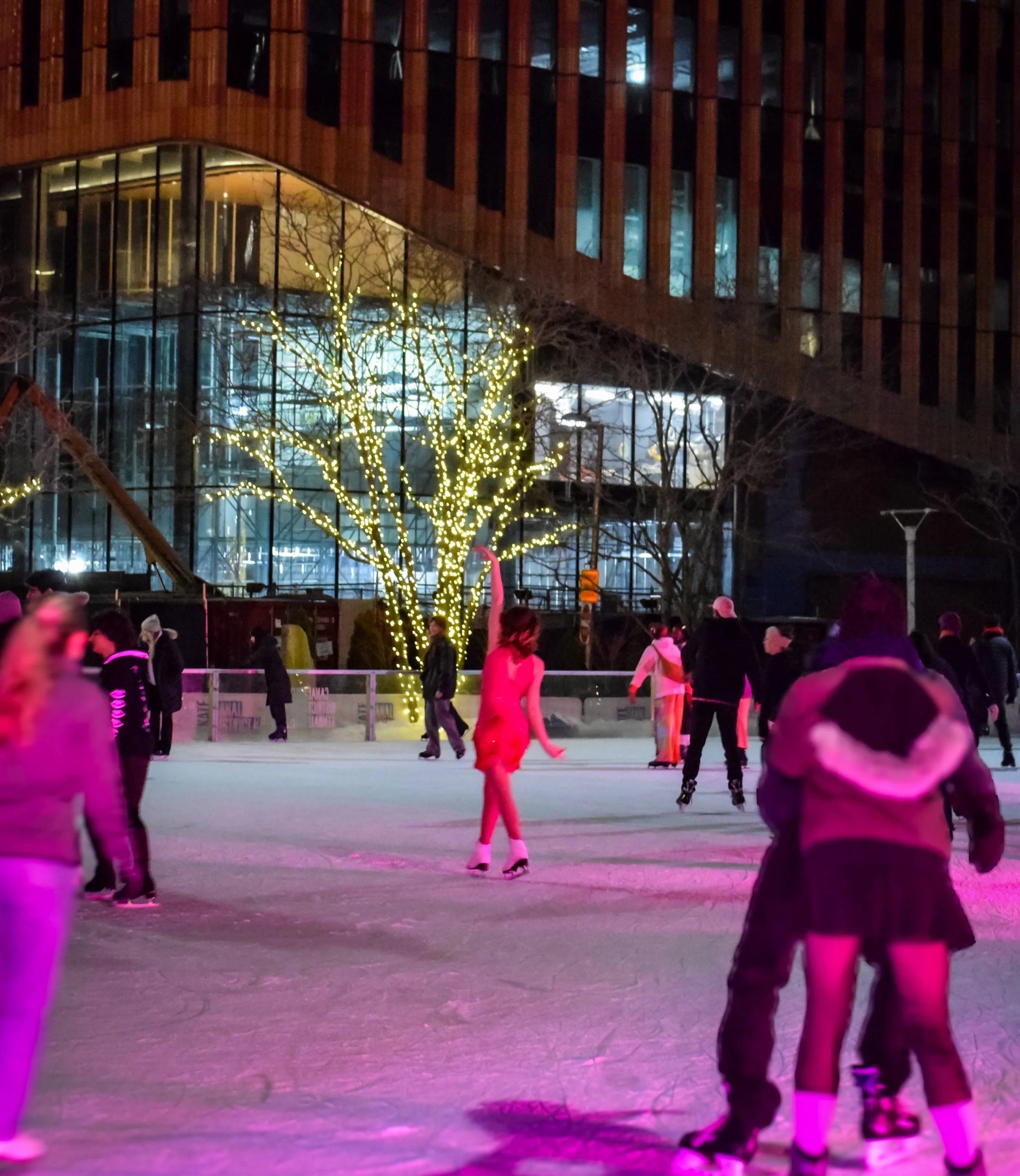 People skating on an ice rink decorated with trees wrapped in string lights, with a modern building in the background at night.