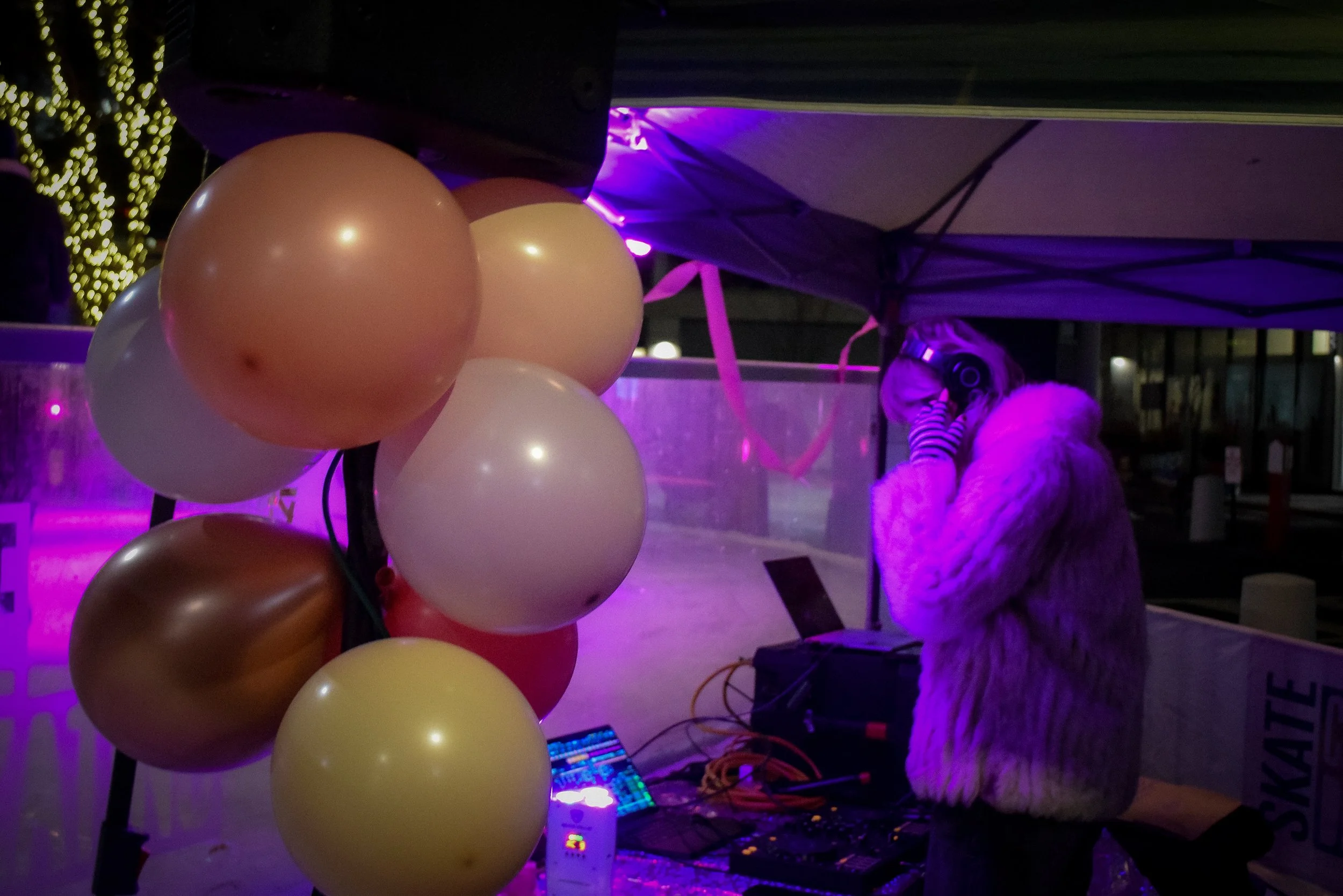 A person with long hair, wearing headphones and a striped shirt, standing behind a DJ setup with a colorful light display, illuminated by purple lighting, near a cluster of balloons in shades of pink, cream, and brown at night.