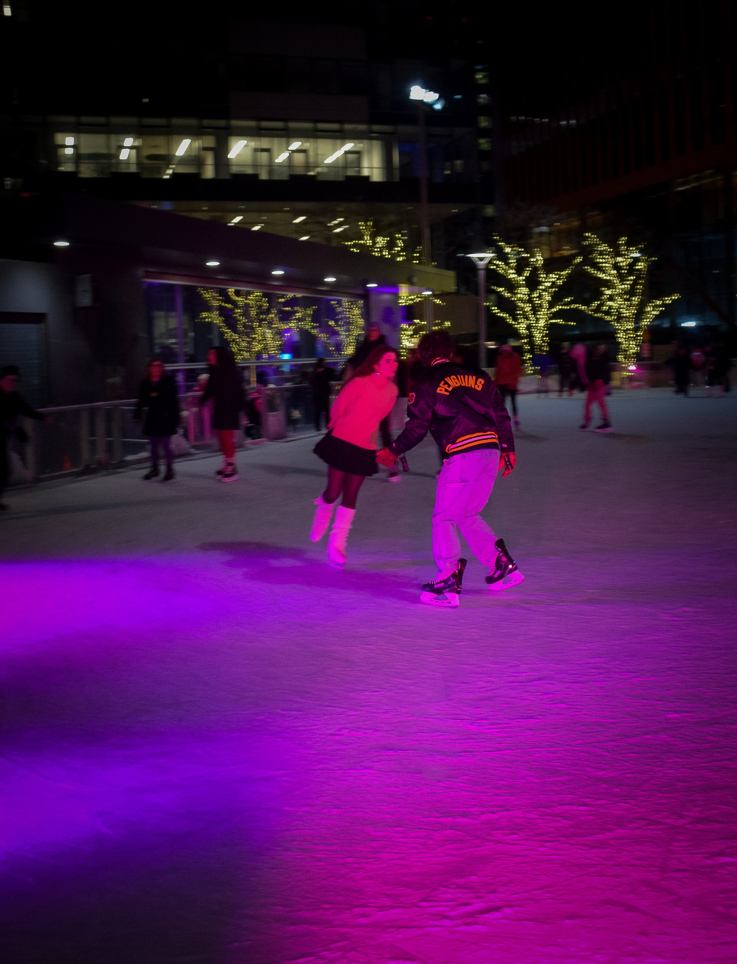 People ice skating at an outdoor rink at night, with city buildings and trees decorated with string lights in the background.