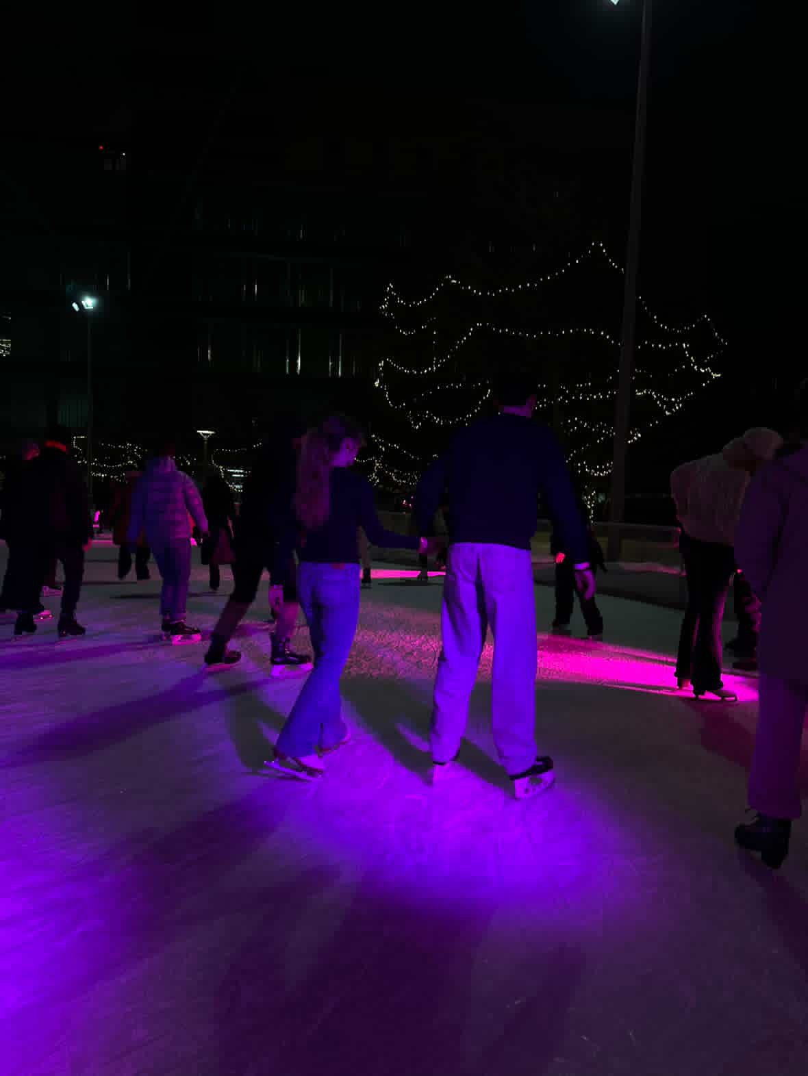 People ice skating at an indoor rink illuminated with colorful purple and pink lights, with string lights hanging above in the background.