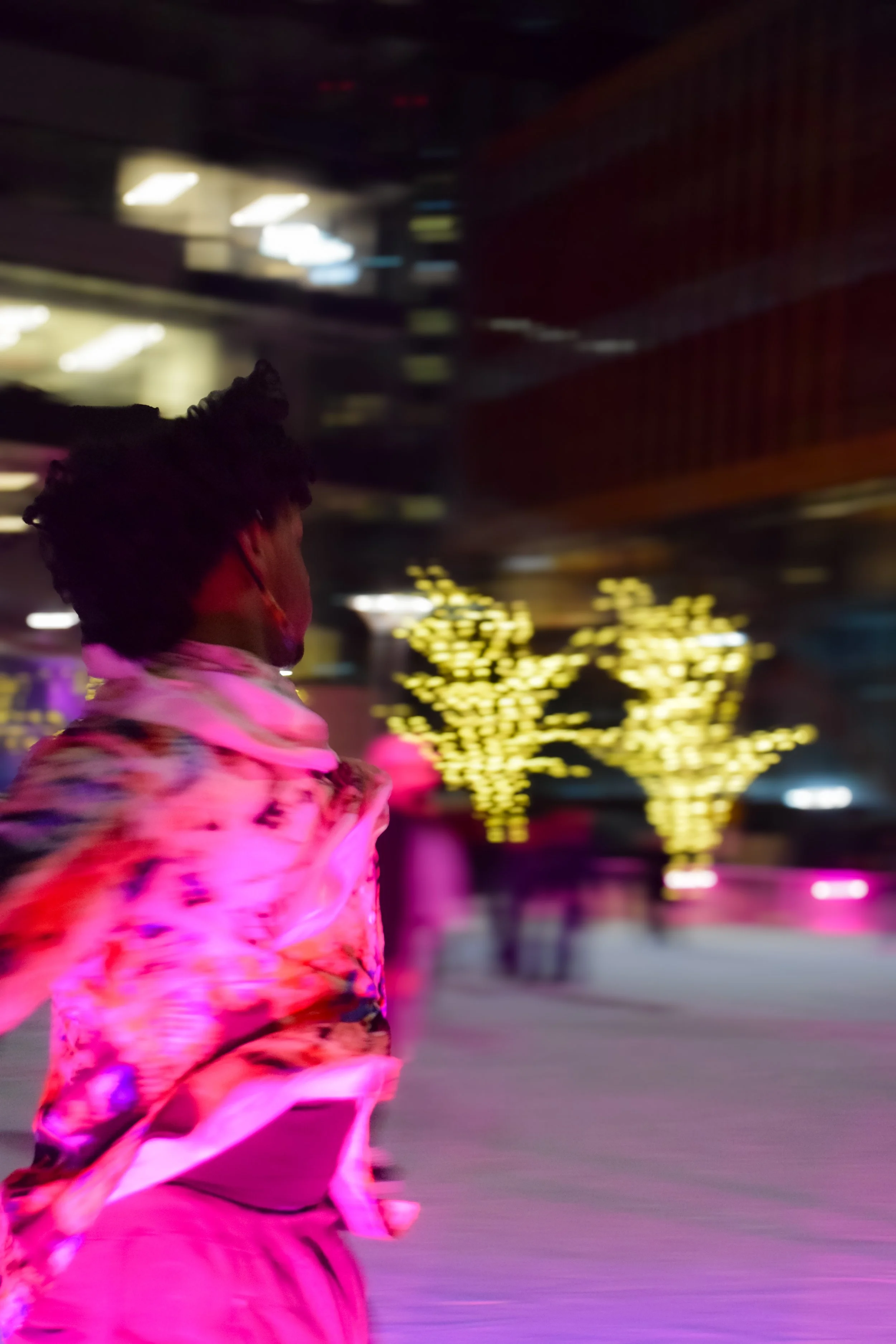 A person with dark, curly hair walking in a dimly lit indoor space decorated with string lights. The person is wearing a vibrant, colorful jacket and a face mask. The background features illuminated buildings or structures with a warm, pinkish glow f
