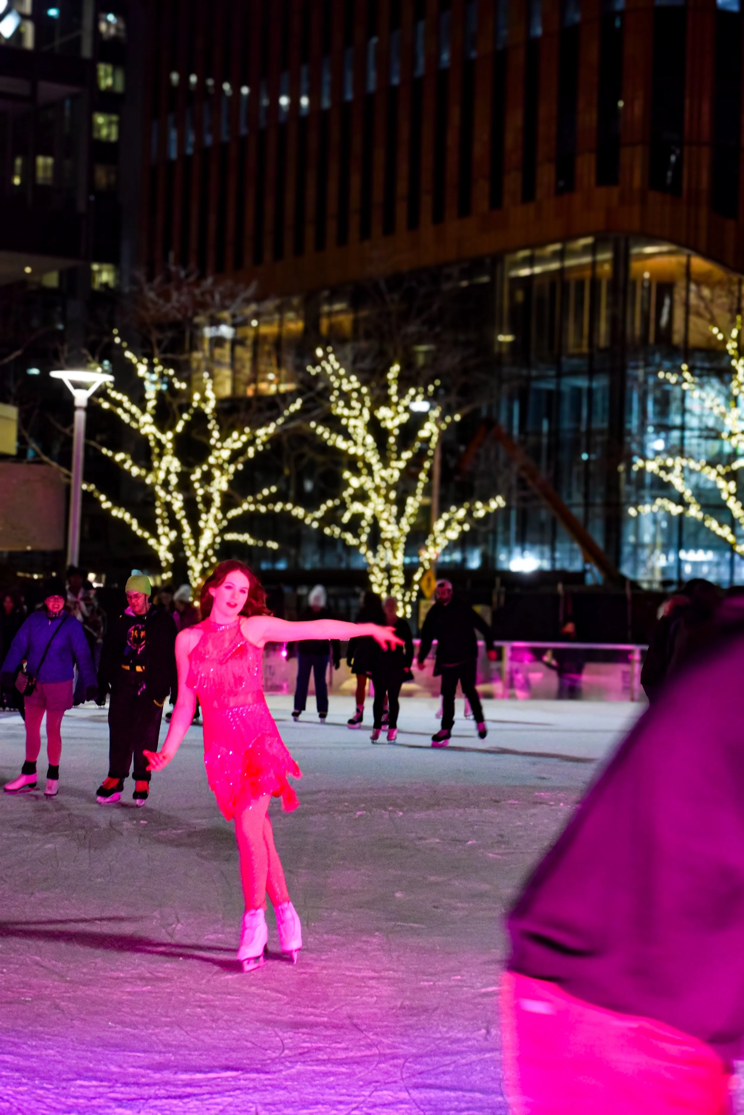 People ice skating outdoors at night with illuminated trees and a modern building in the background.