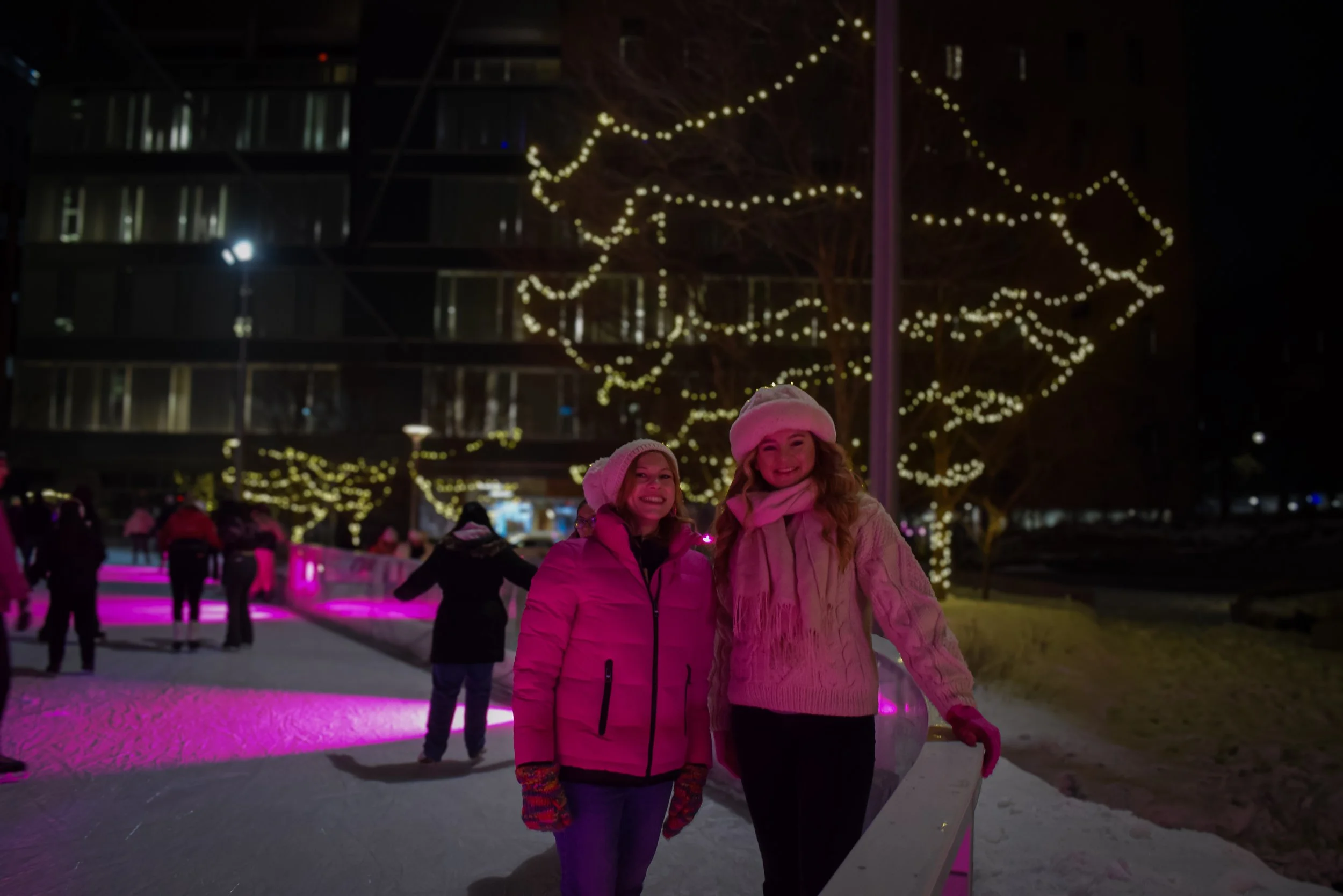 Two women in winter clothing and hats smiling at the camera on an outdoor ice skating rink at night, illuminated by festive string lights hung above.