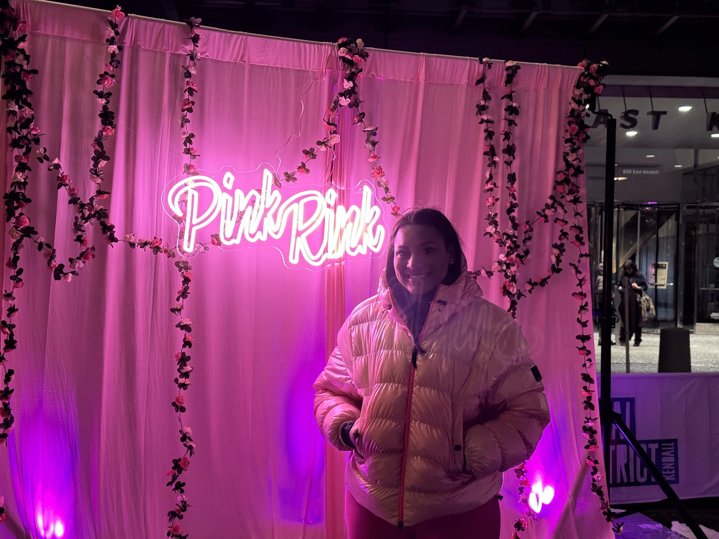 A woman wearing a pink puffer jacket stands in front of a pink curtain decorated with artificial flowers, illuminated by pink neon lights that spell 'Pink'.