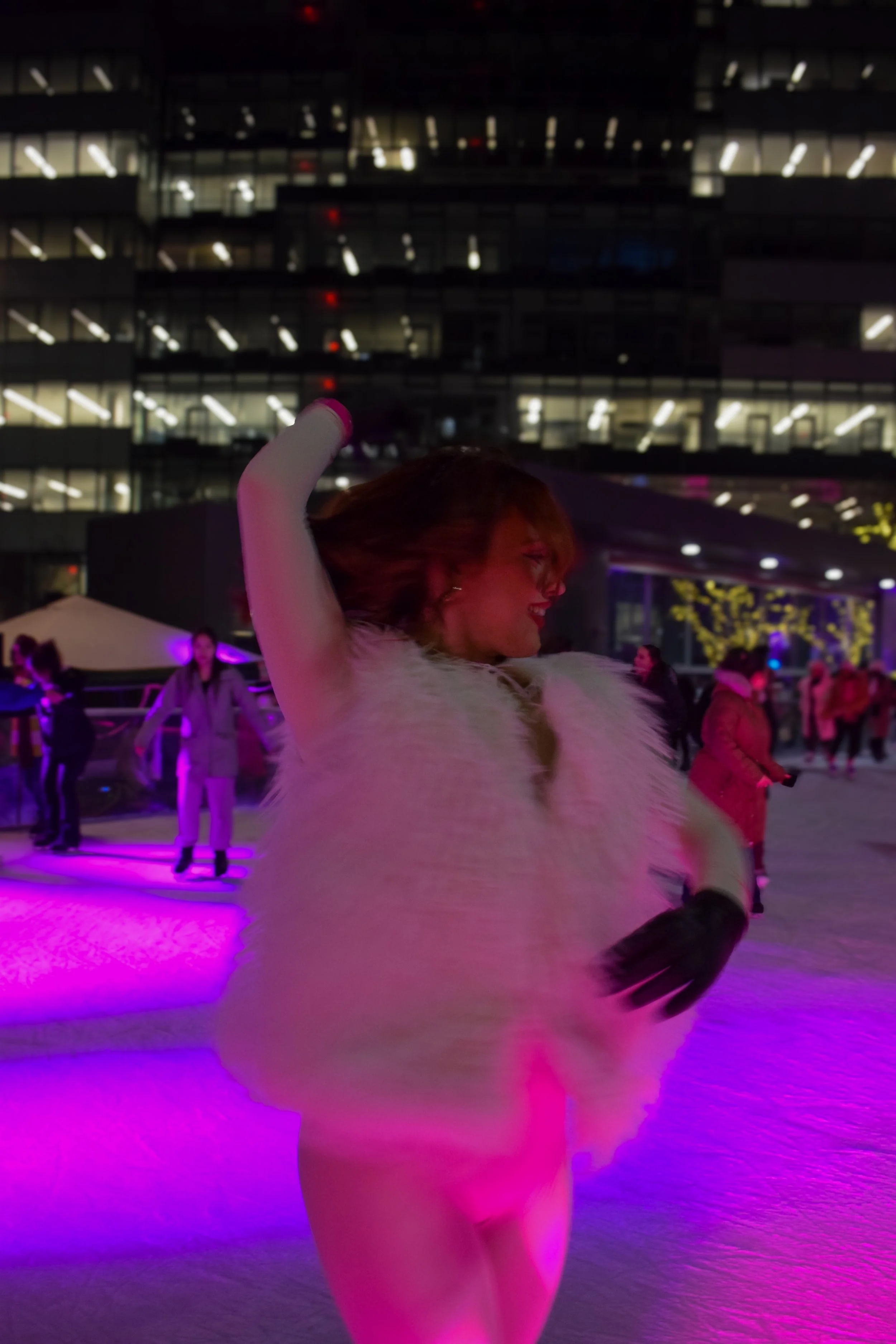 A woman ice skating at an outdoor rink at night, smiling and raising her arm, with illuminated buildings and other ice skaters in the background.