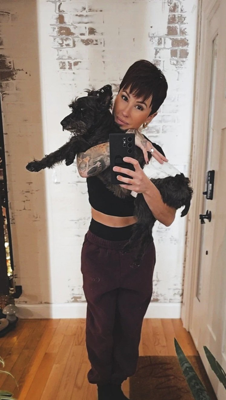 A woman with short dark hair and tattoos on her arm taking a mirror selfie with a large black dog in her arms in a room with a white brick wall and hardwood floor.